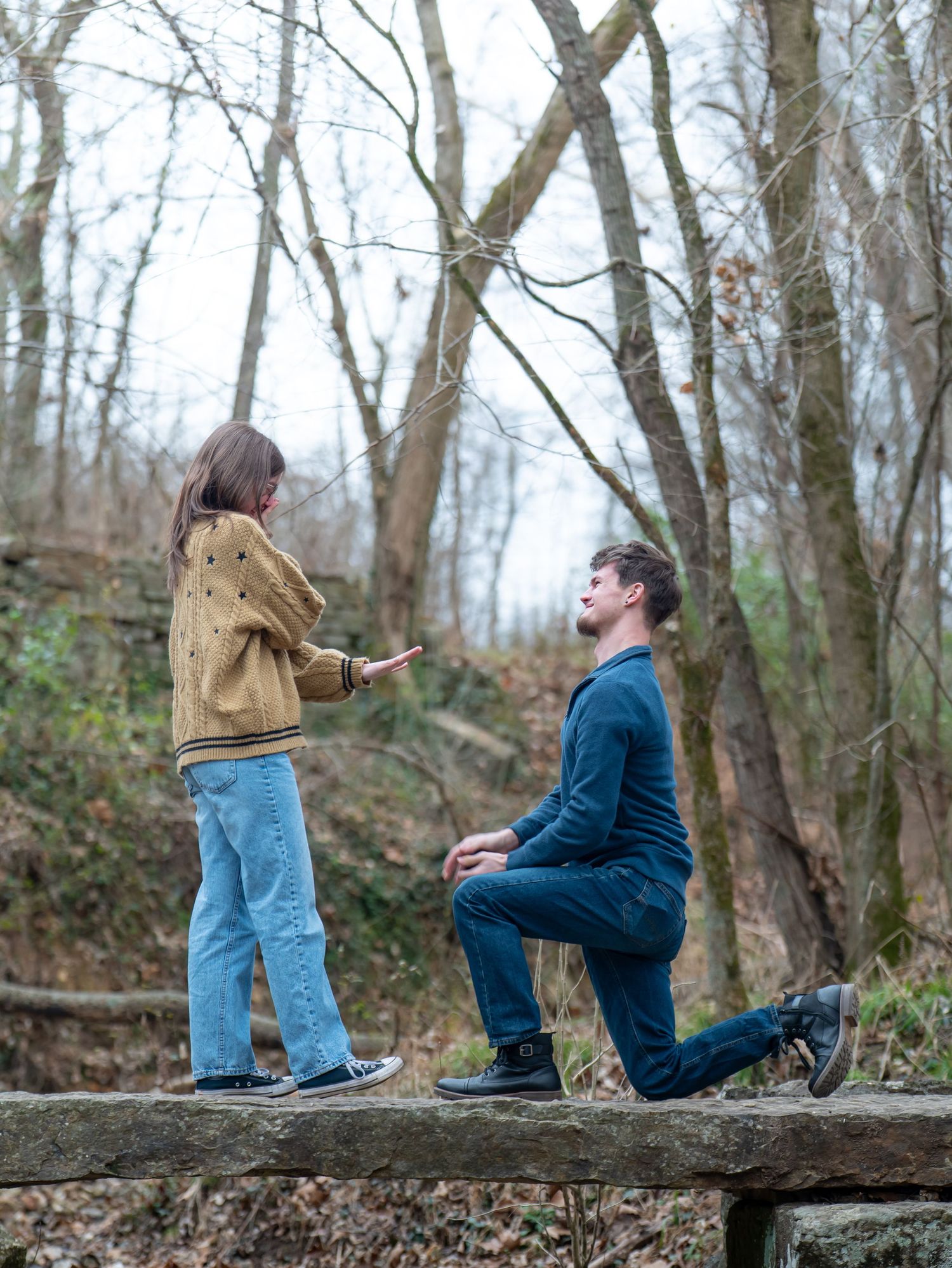 A couple shares a special moment on a fallen log in a winter forest during what appears to be a proposal.