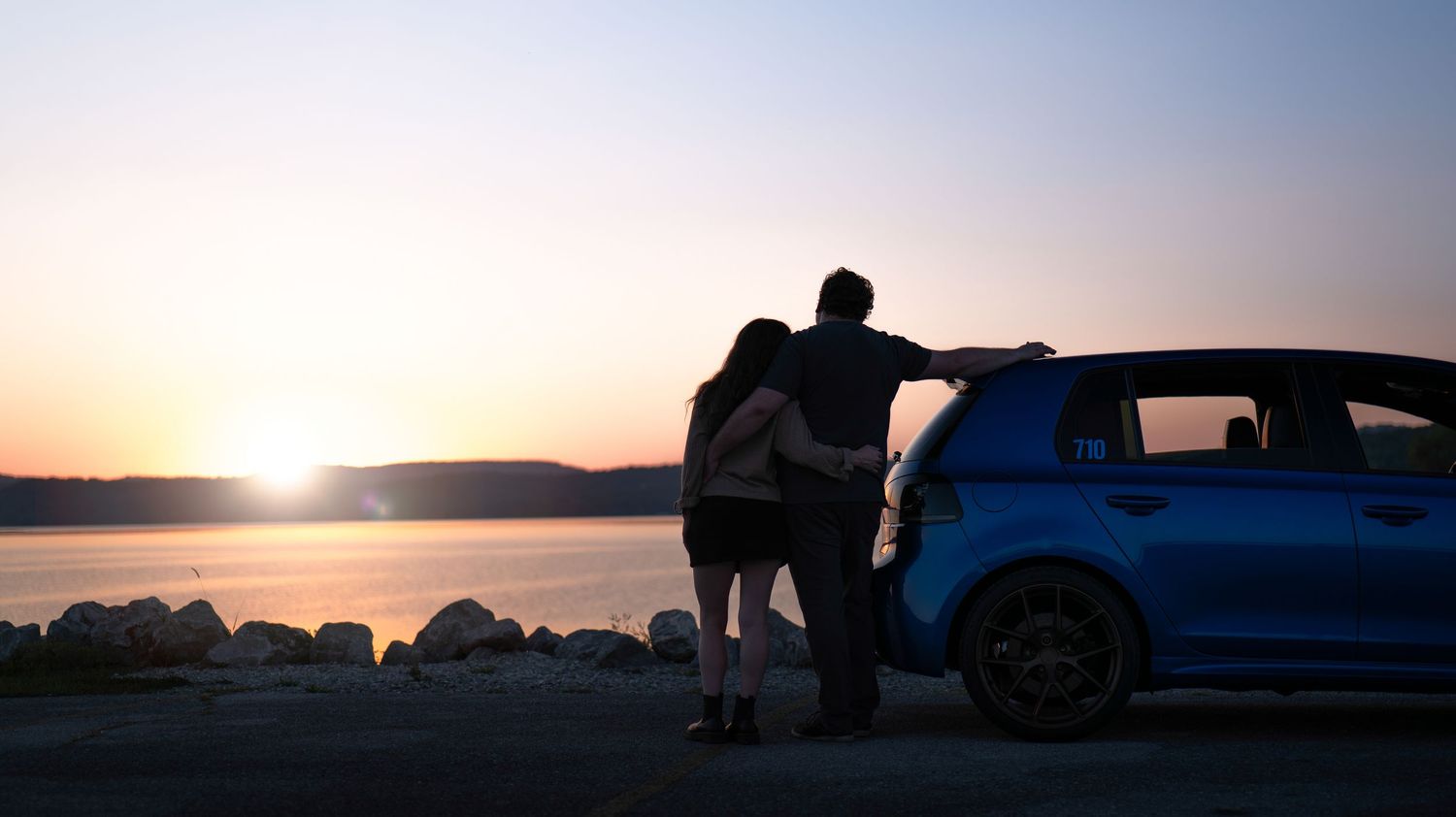Silhouette of people leaning against a car watching sunset over a lake.