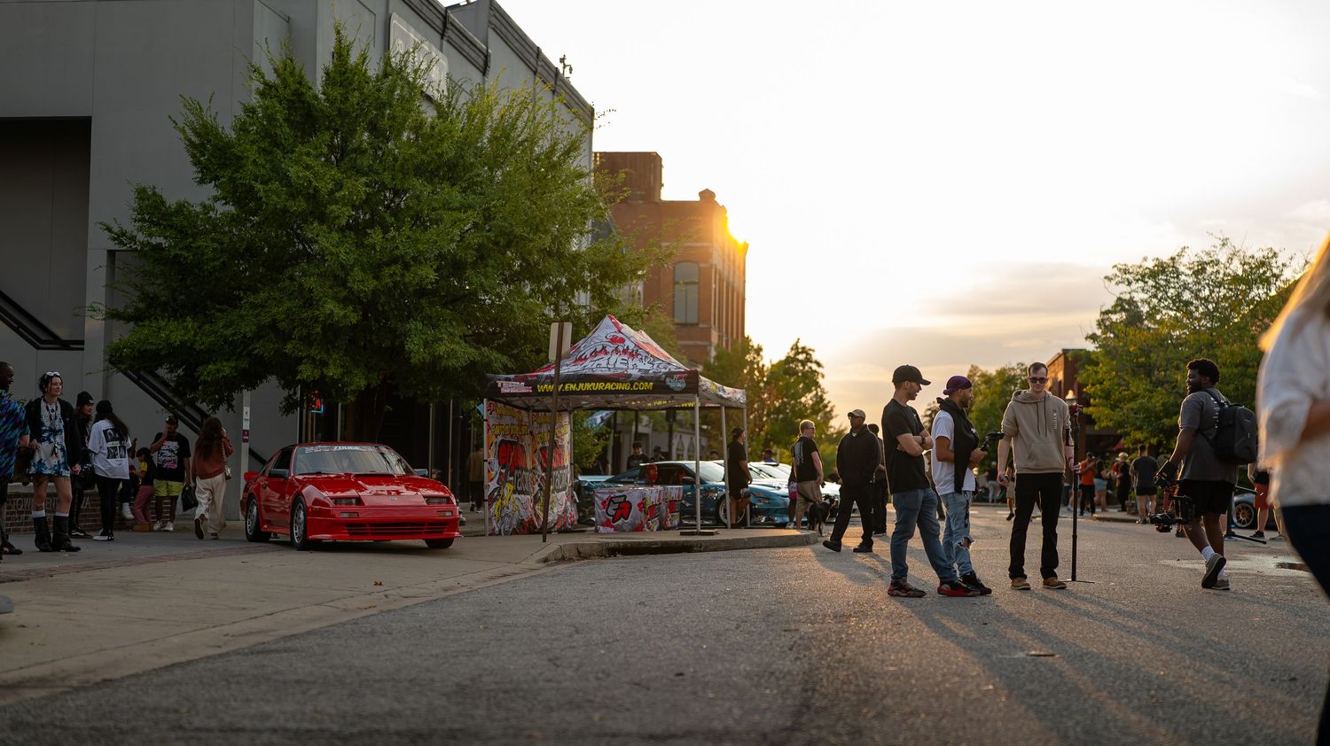 Wide panoramic view of a car meet event with buildings and trees at dusk.