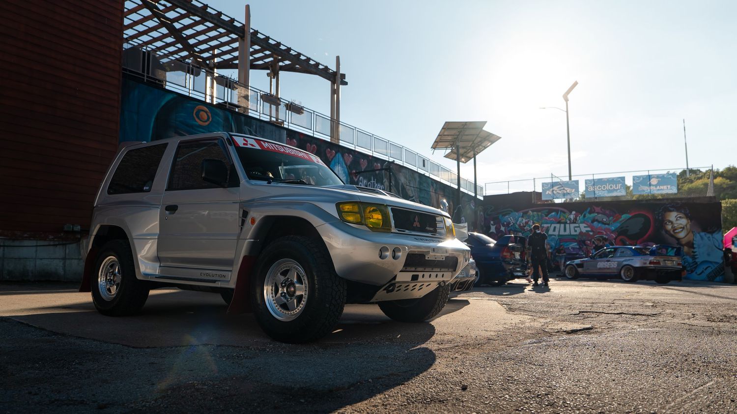 Wide angle shot of vehicles parked in front of a graffiti-covered wall under a blue sky.