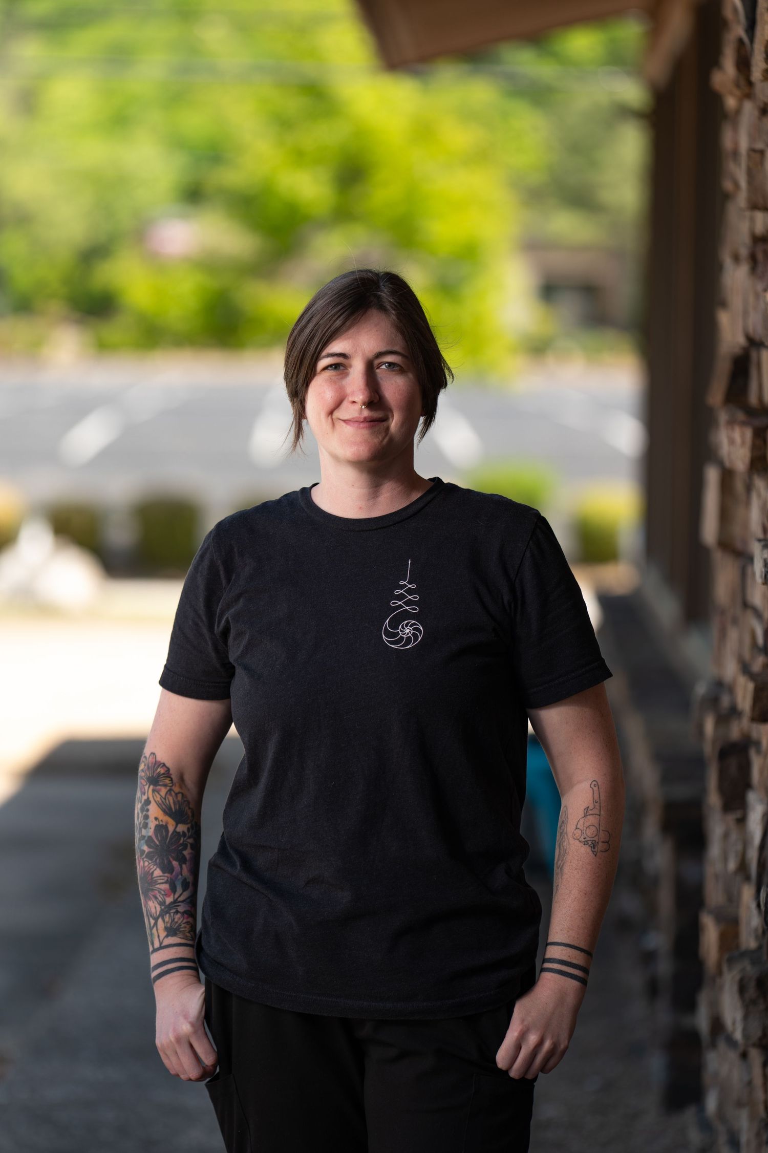 Smiling woman with tattoos wearing a black graphic t-shirt standing outdoors with green trees in the background.