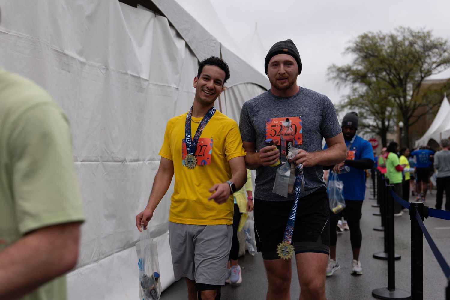 Two marathon runners smiling and posing together after a race, wearing medals and athletic gear outdoors.
