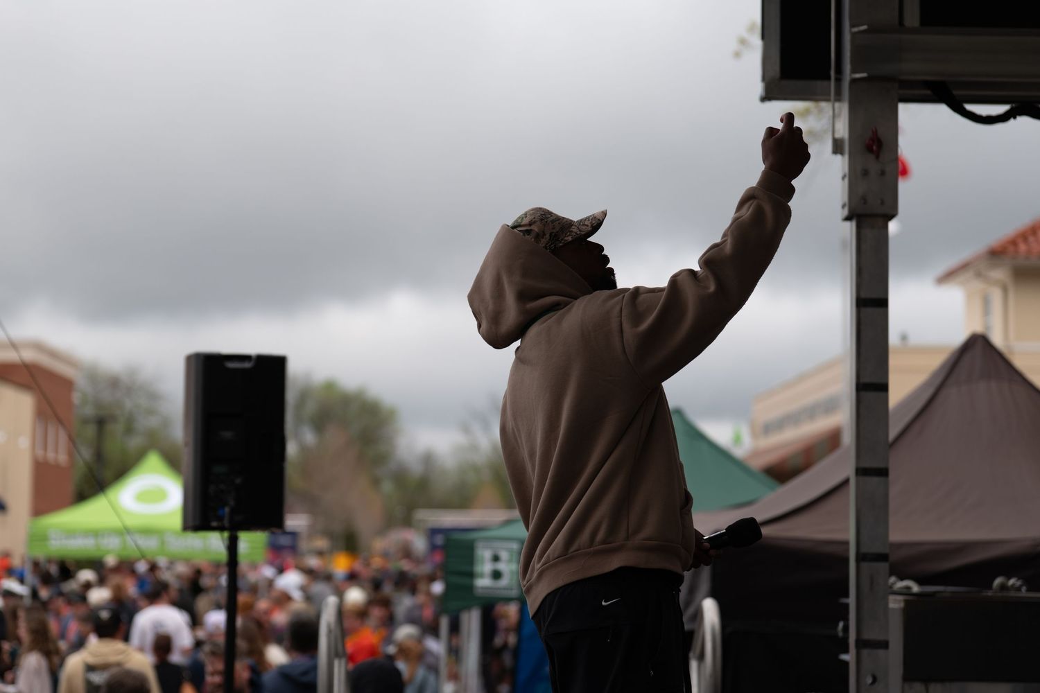 Man in hoodie raises fist on stage at outdoor festival with crowd and green tent in background.