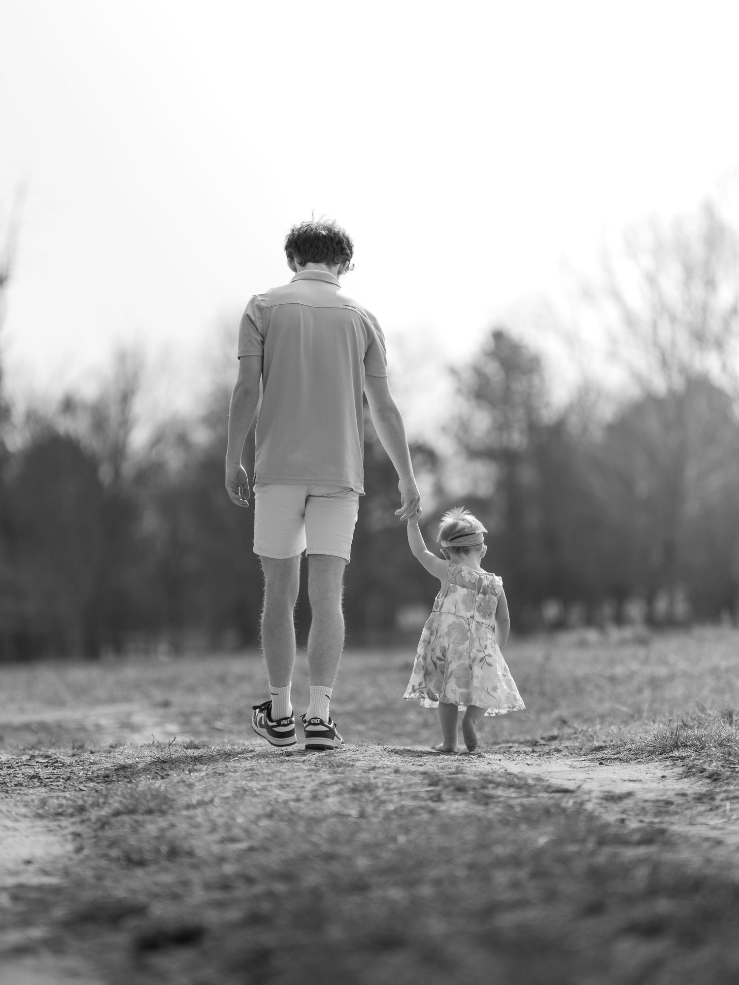Black and white photo of a father holding hands with his toddler daughter walking down a path in a park.