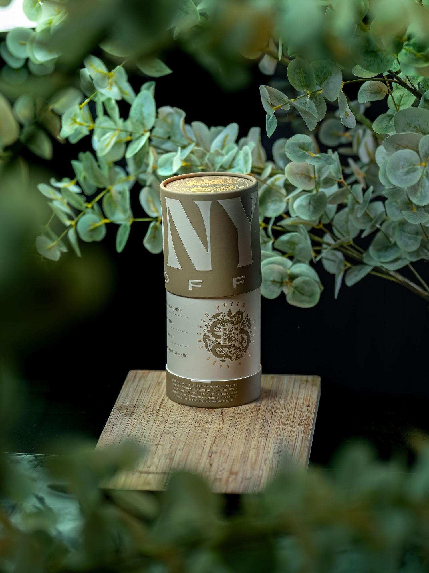 Cylindrical eco-friendly product packaging with gold botanical branding displayed on wood block surrounded by lush green eucalyptus leaves.