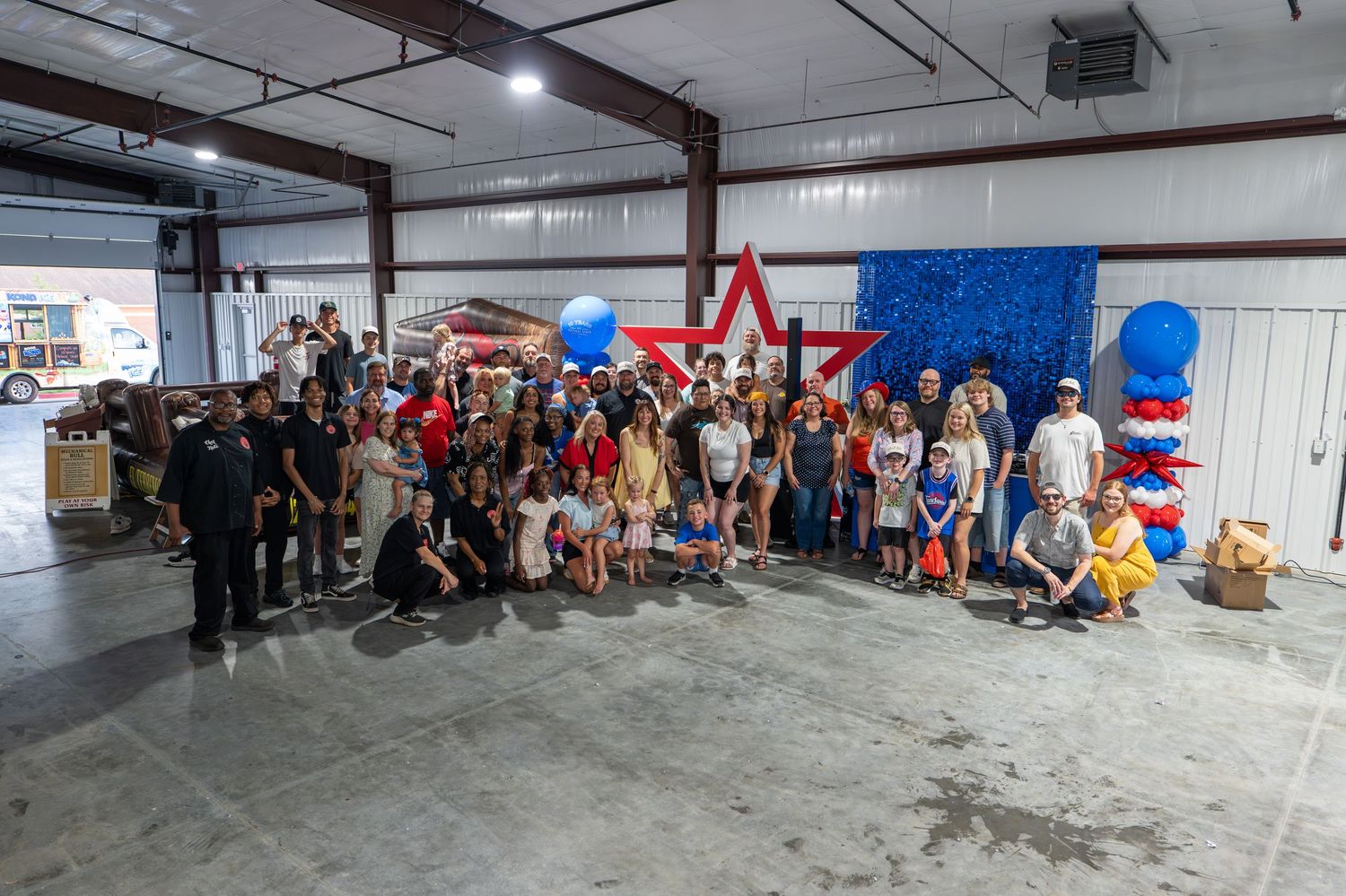 Large group photo inside a decorated warehouse with patriotic red, white, and blue balloons and star backdrop.