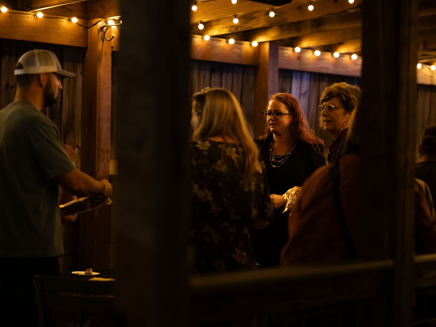 People gathered in a dimly lit venue with string lights overhead having a conversation in warm ambient lighting.