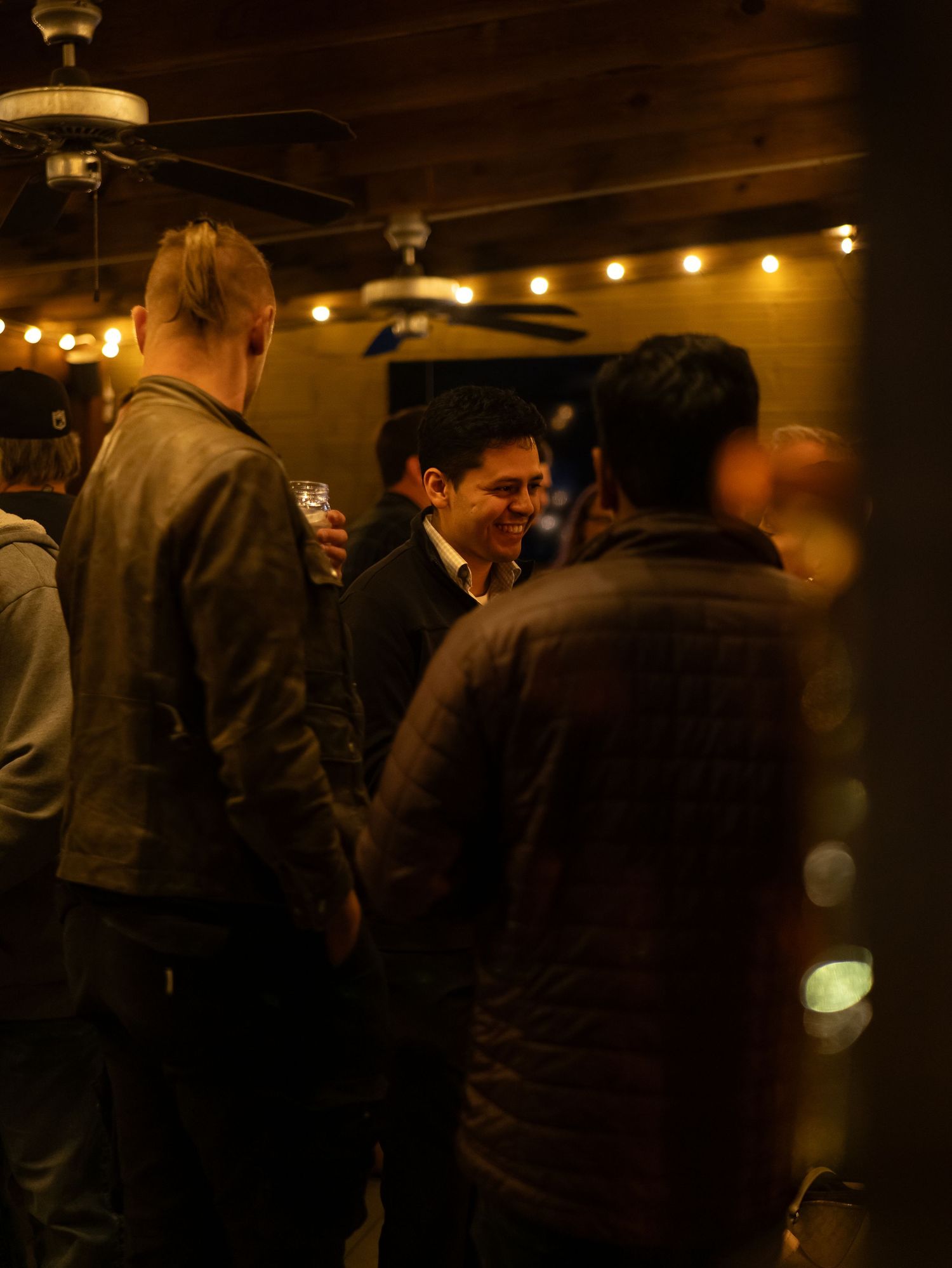 People gathered at a dimly lit indoor venue with warm lighting and string lights creating an intimate atmosphere.