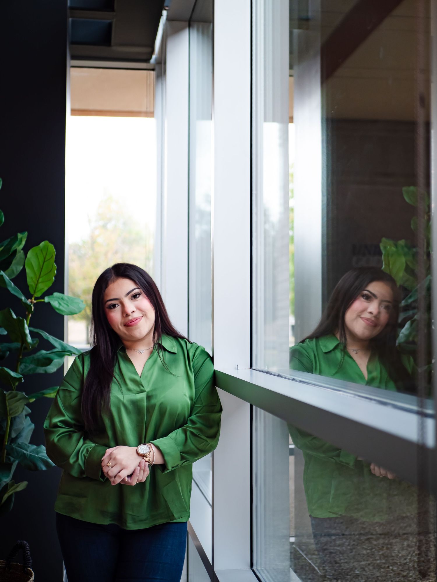 Person in a green hoodie poses by a window with natural light and plants in the background.
