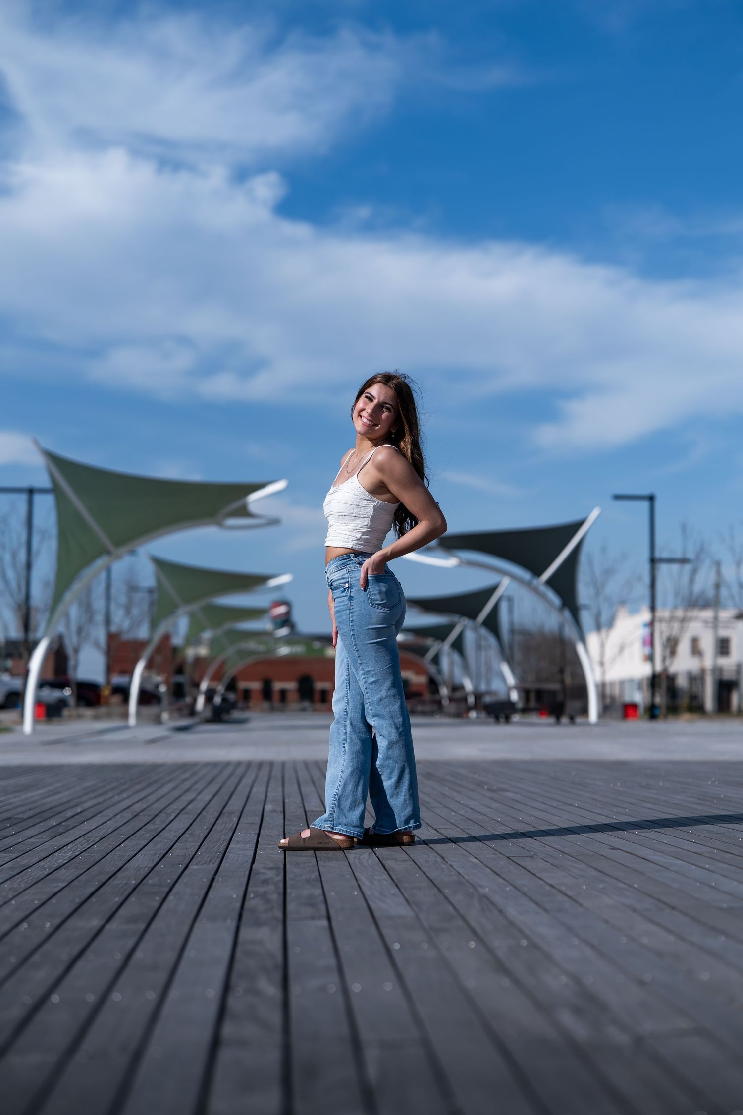 Young woman in white crop top and wide-leg jeans posing on wooden deck under blue sky with green shade sails.