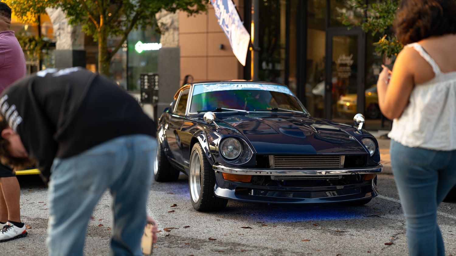 Custom lowered cars displayed at an outdoor car show with autumn trees overhead.