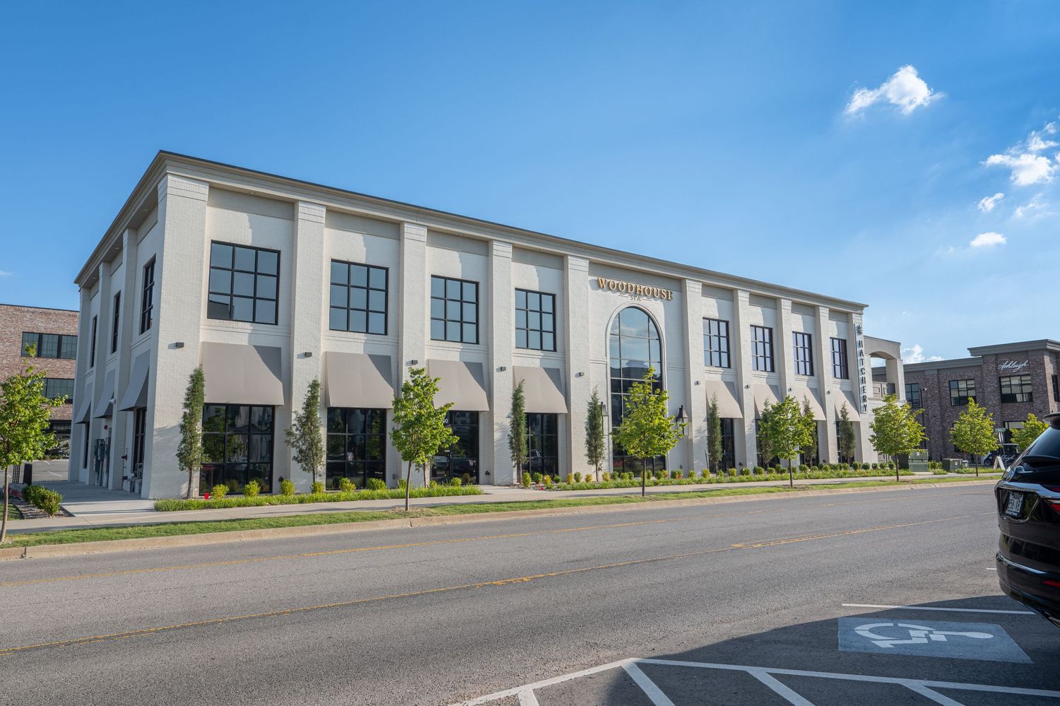 Modern multi-story commercial building with large arched windows, white facade, and landscaped sidewalk under a clear blue sky.