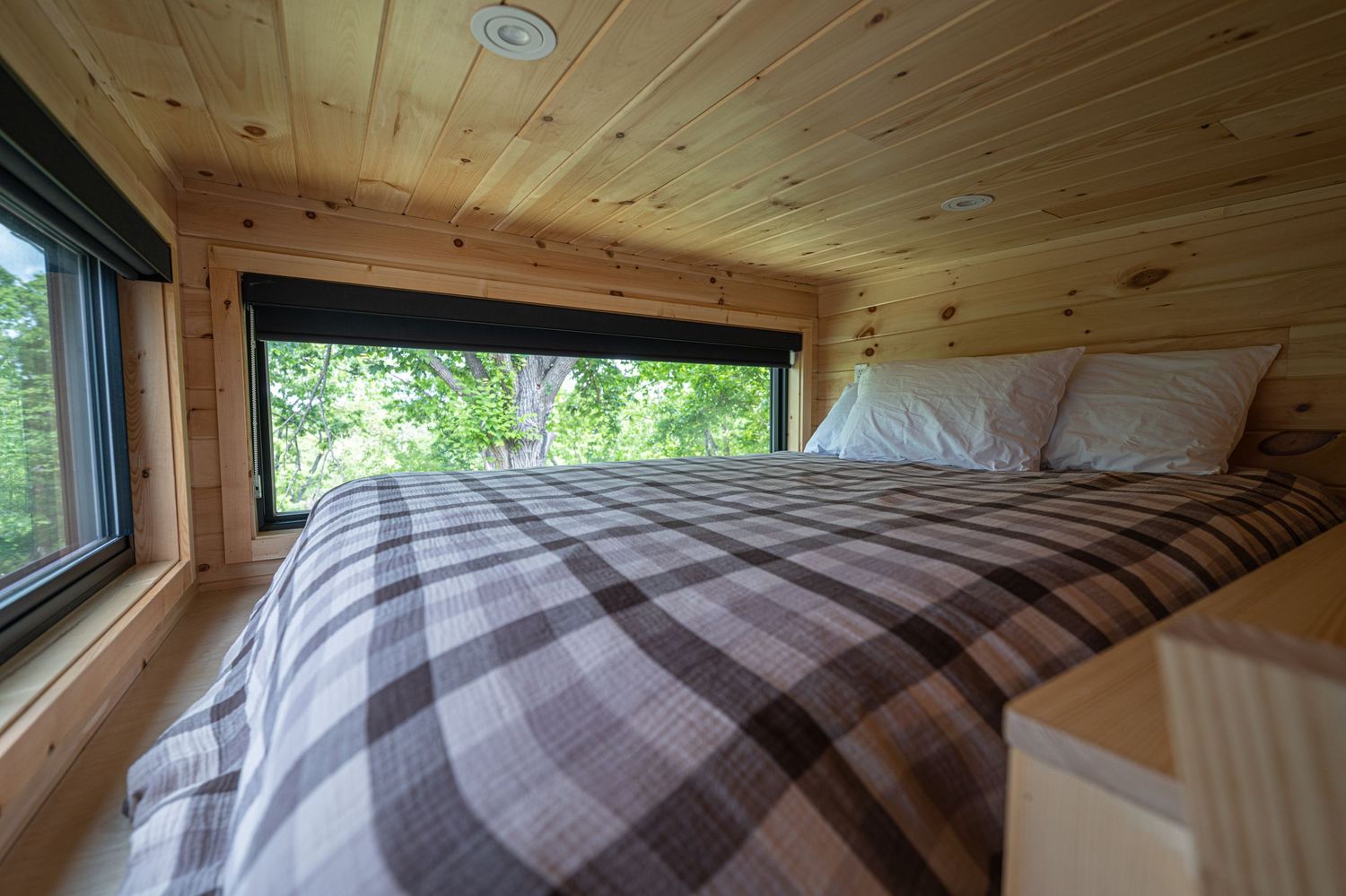 Cozy tiny home loft bedroom with plaid bedding, wood-paneled ceiling, and large window overlooking lush green trees.