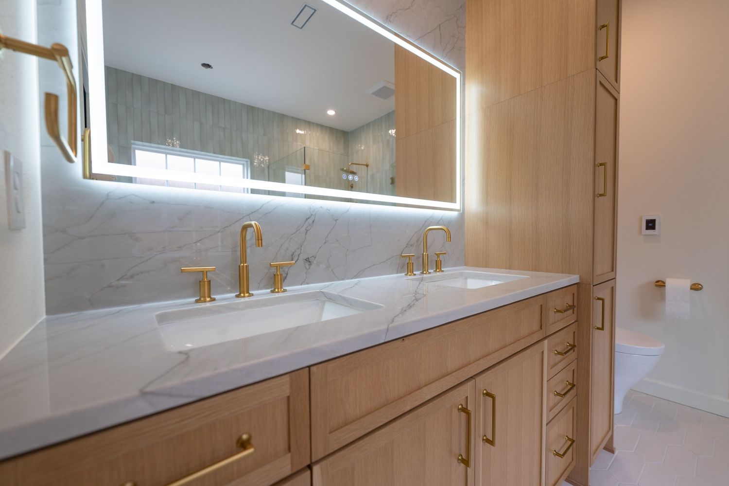 Modern bathroom with dual gold faucets, marble countertop, wood vanity, and backlit LED mirror.