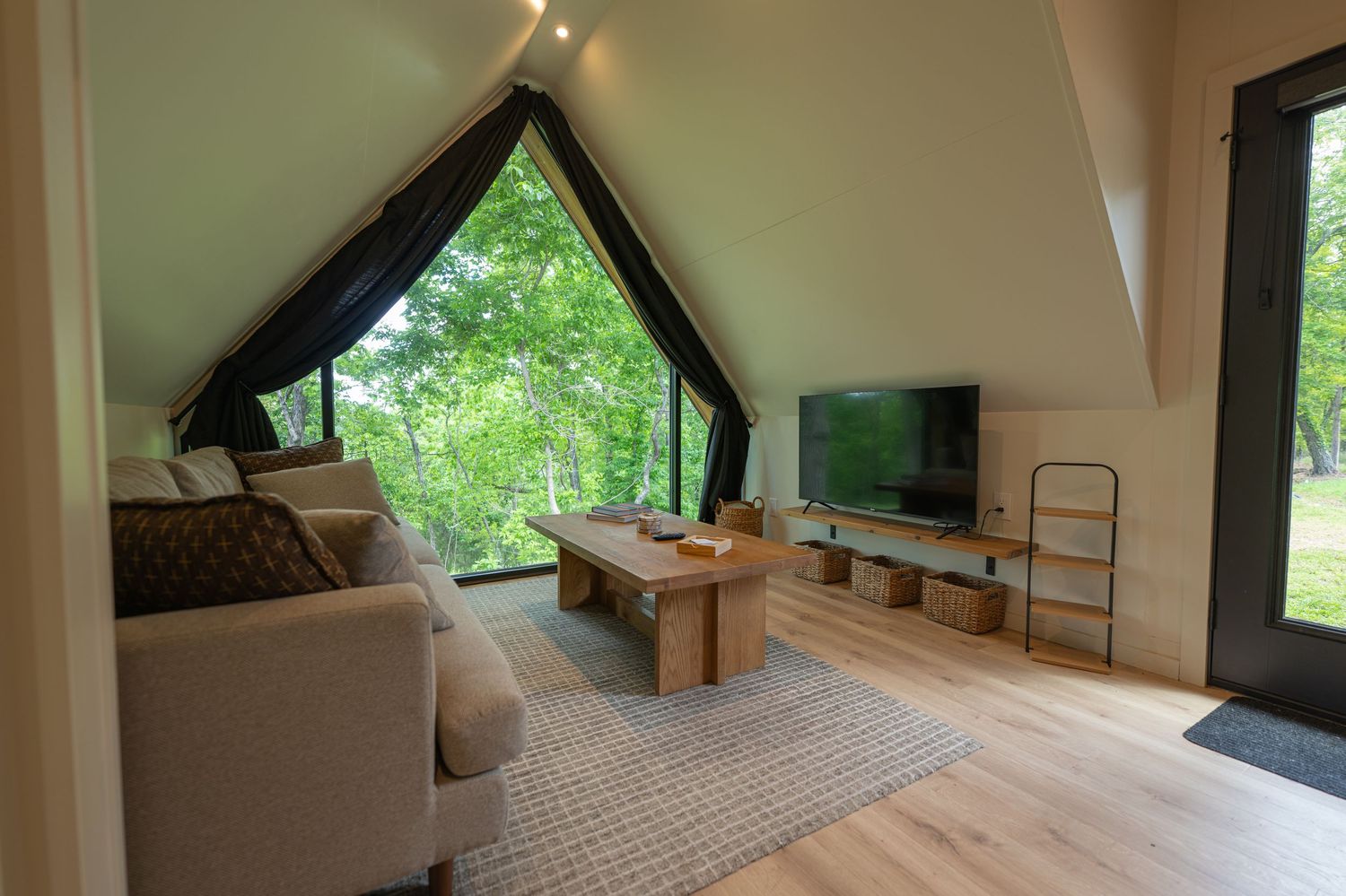 A-frame cabin living room with large triangular window overlooking trees, featuring a sofa, coffee table, and flat-screen TV.