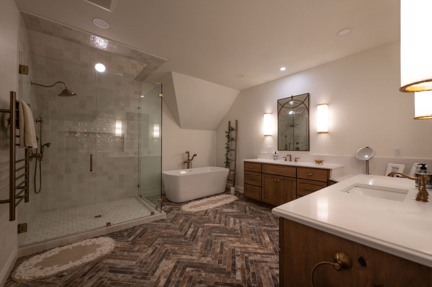 Luxury bathroom with herringbone brick floors, glass shower, freestanding tub, and dual vanity under vaulted ceiling.