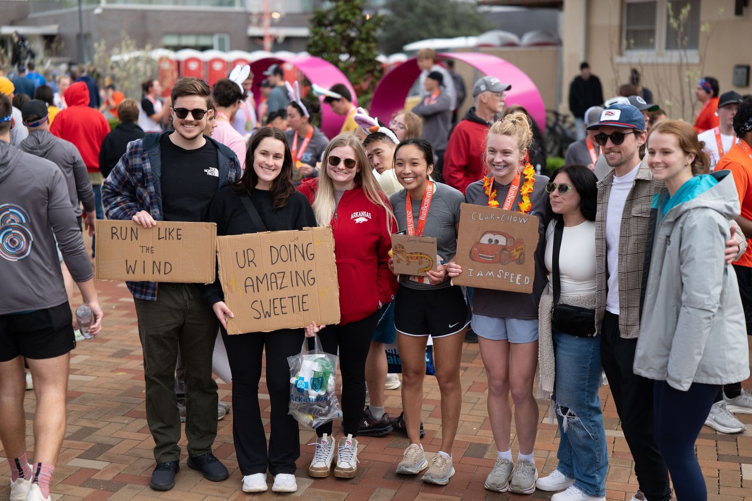 Group of friends holding encouraging cardboard signs cheering on marathon runners at an outdoor race event.