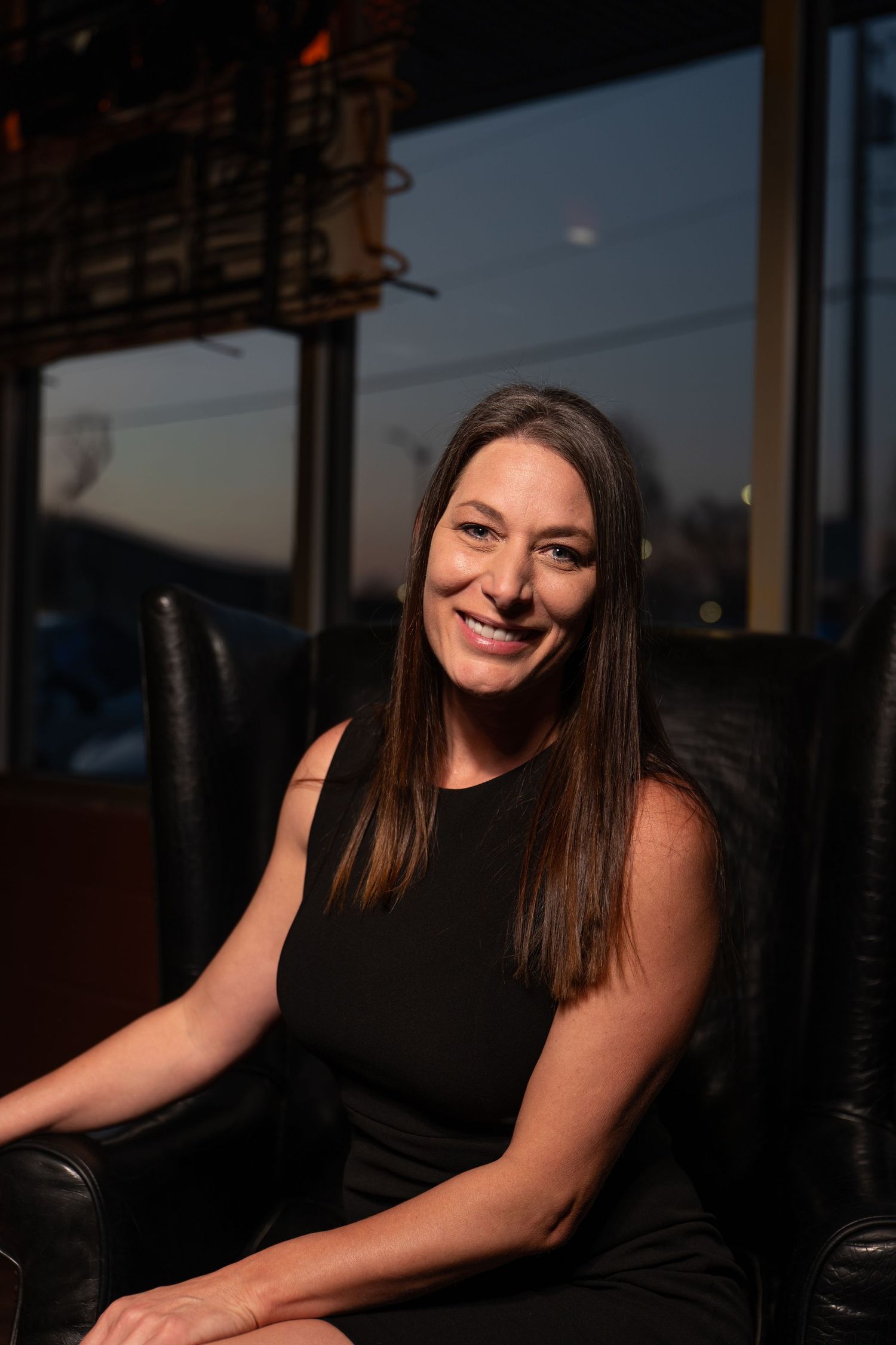 Smiling brunette woman in black sleeveless dress seated in leather chair at upscale restaurant.