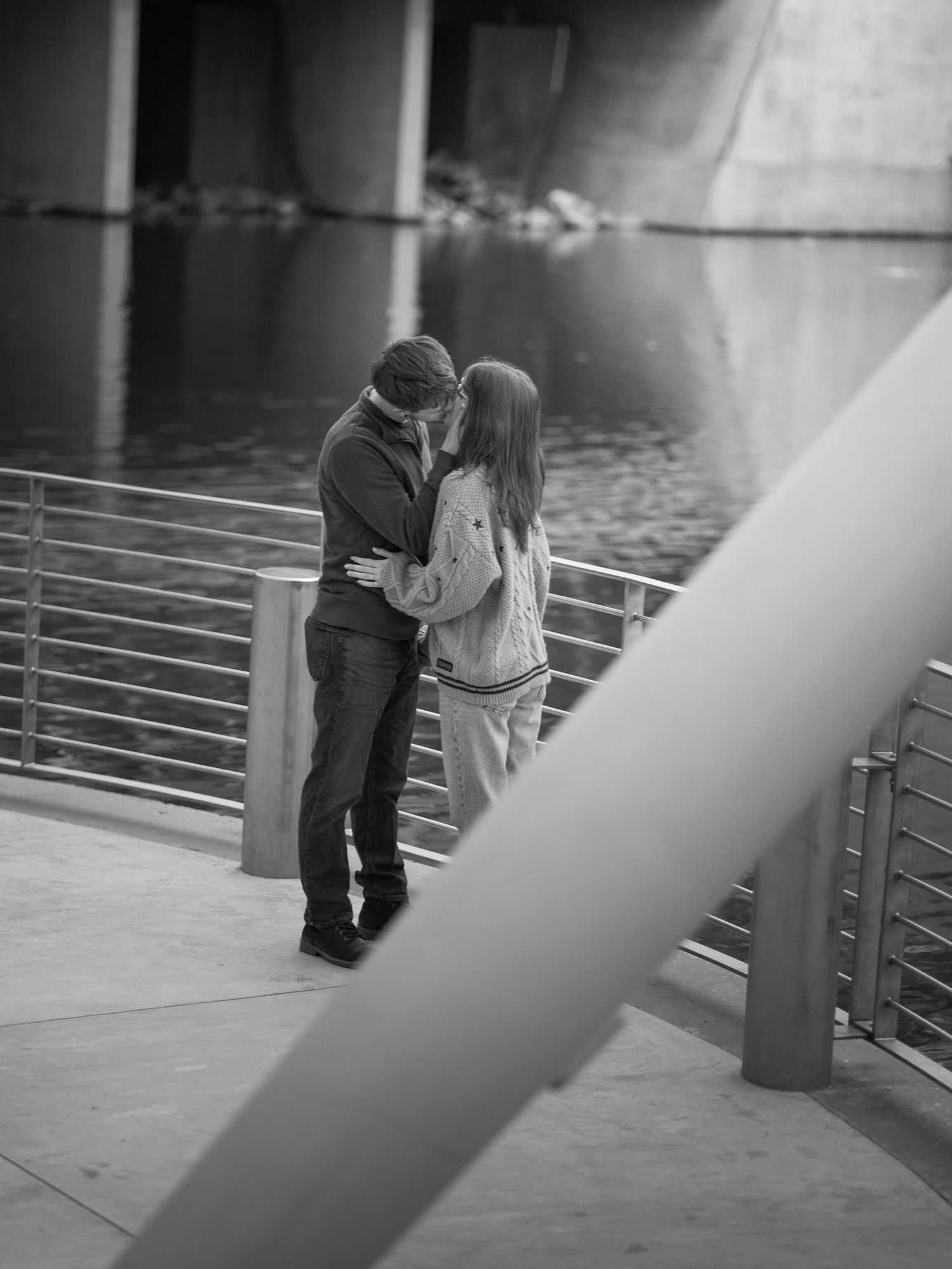 A romantic black and white photo of a couple sharing an intimate moment on a waterfront deck with metal railings.