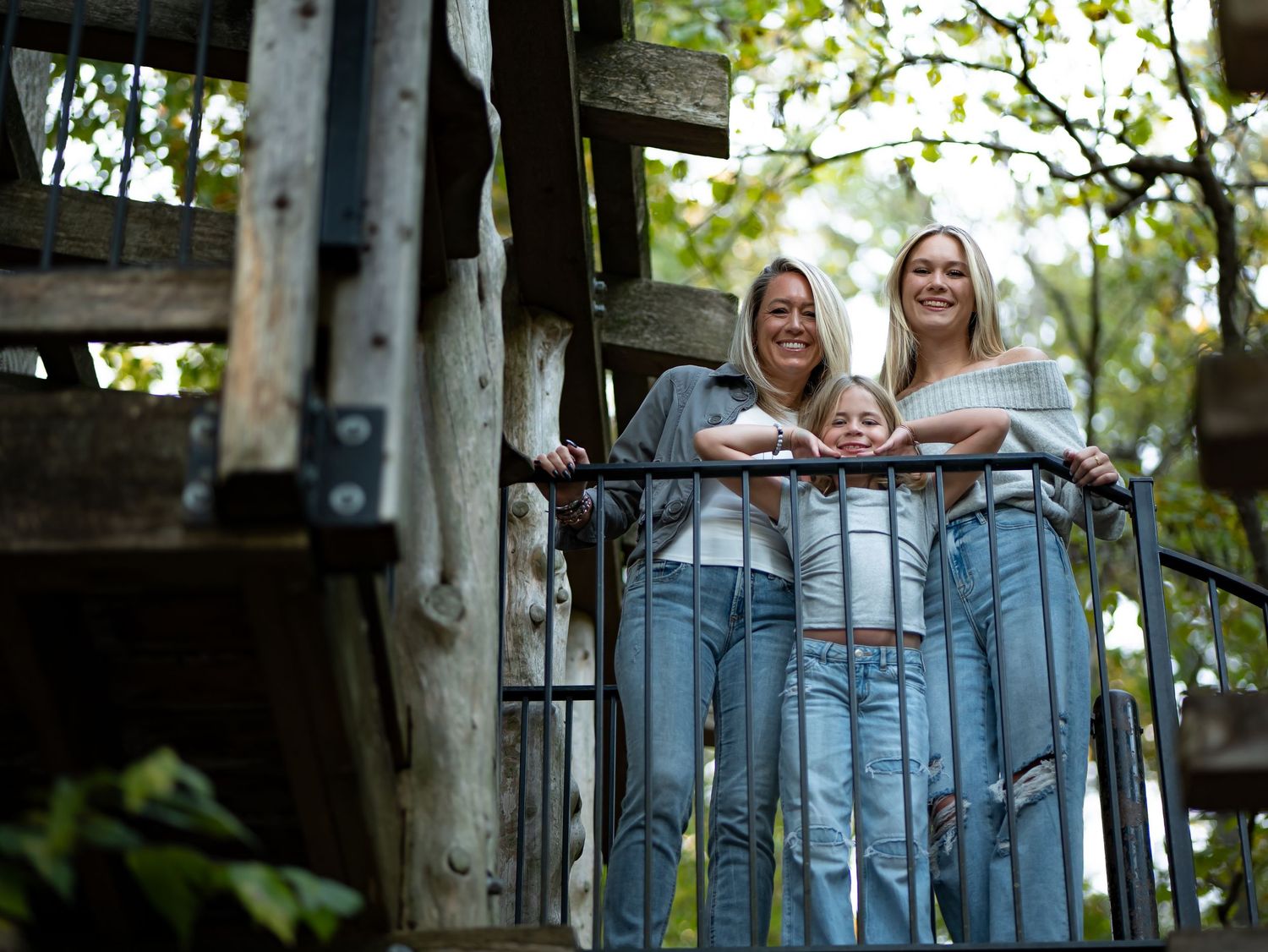 Two people in denim jeans lean against a wooden railing outdoors in a treehouse-like setting.