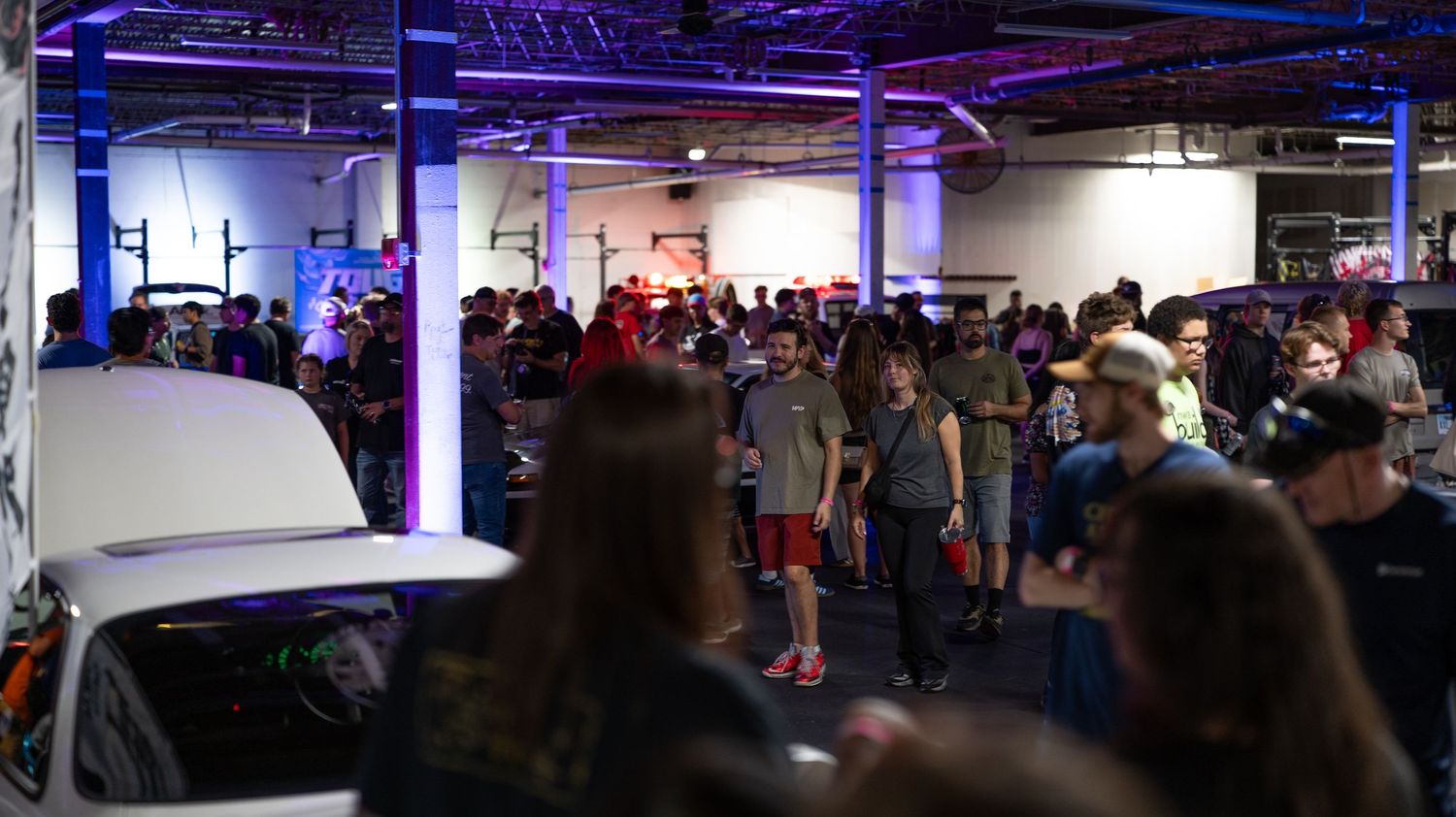 Large crowd gathered at an indoor car show with purple lighting and white vehicle on display.