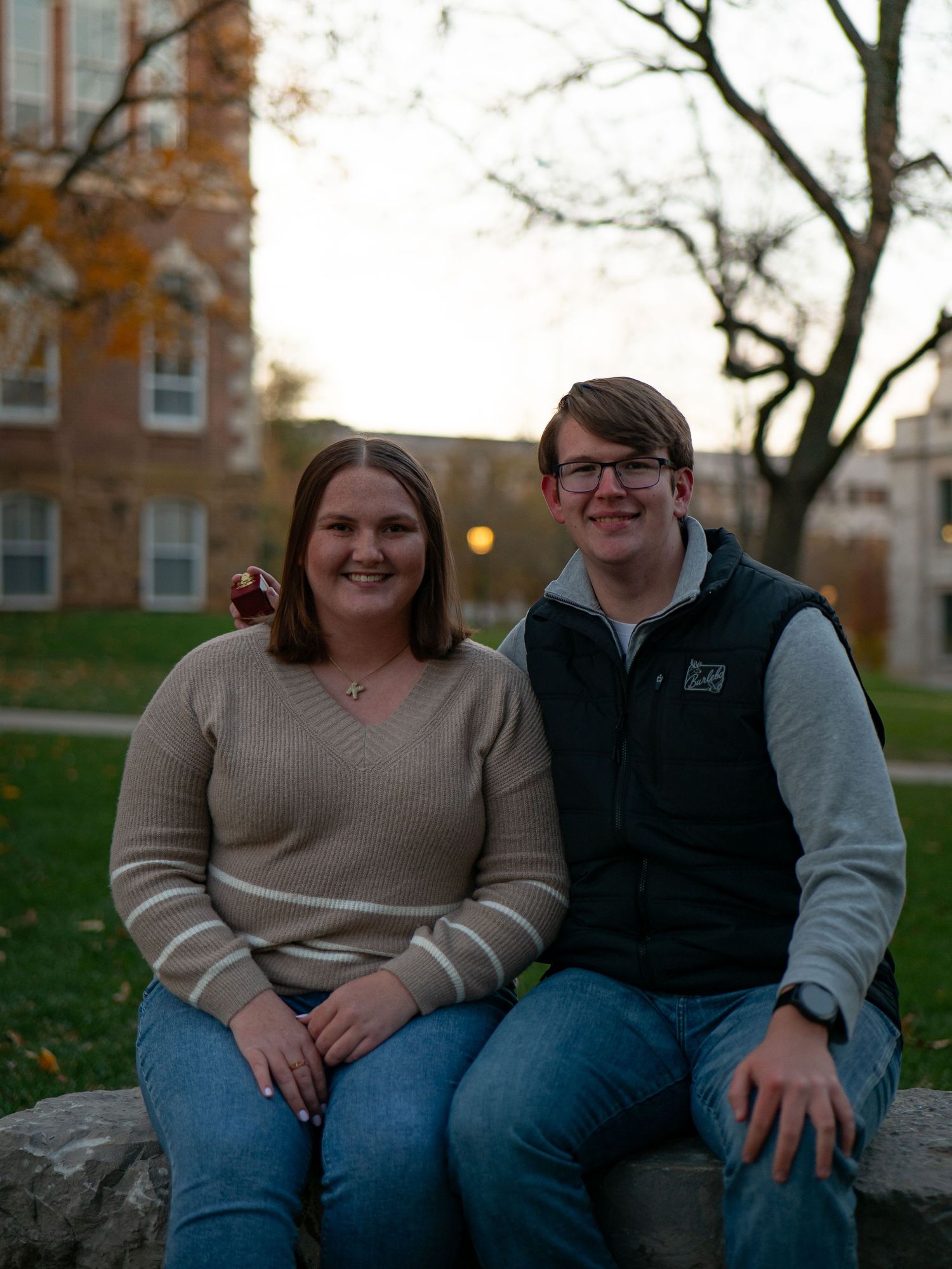 A couple sits together outdoors on a stone wall during an autumn day wearing casual attire.