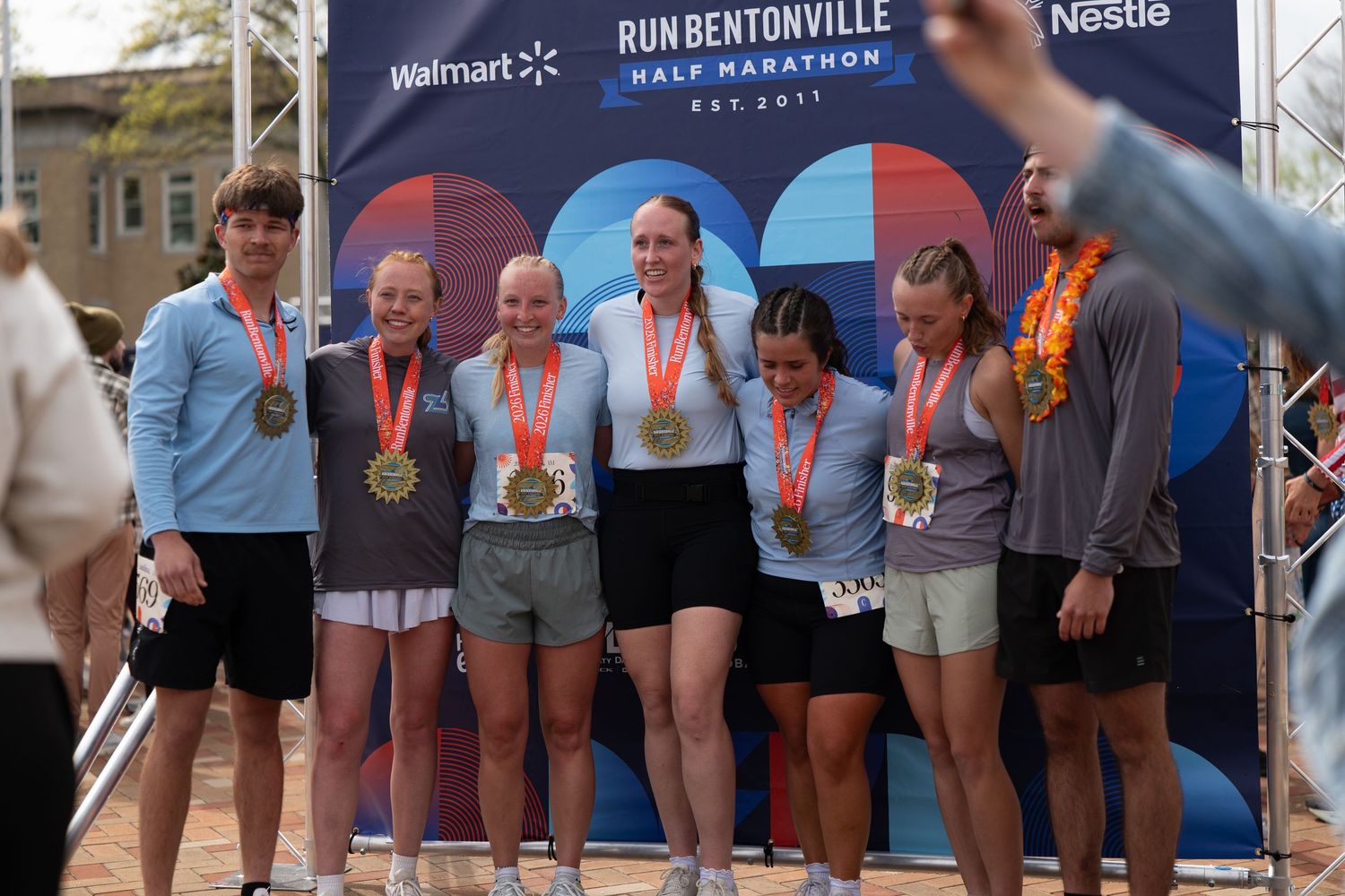 Group of runners wearing gold medals pose at Run Bentonville Half Marathon finish line banner with Walmart and Nestle logos.