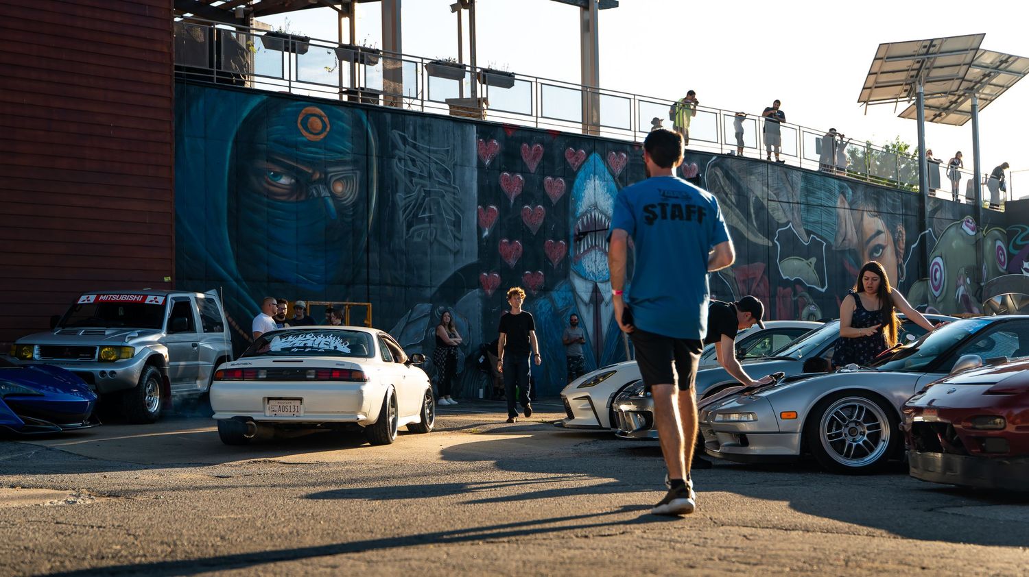 Car enthusiasts line up against a brick wall backdrop at an evening car meet.