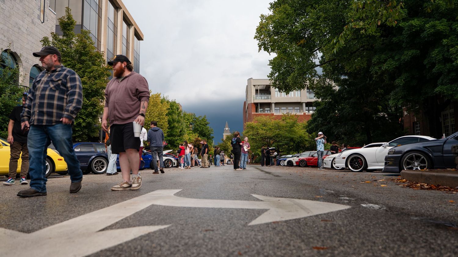 Panoramic street view showing parked cars and pedestrians along a tree-lined urban area.