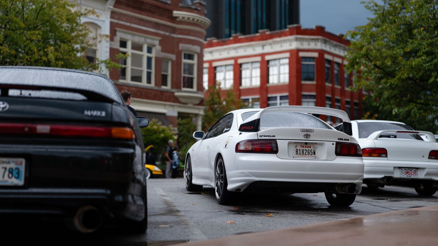 Row of custom cars parked along brick building during automotive event.