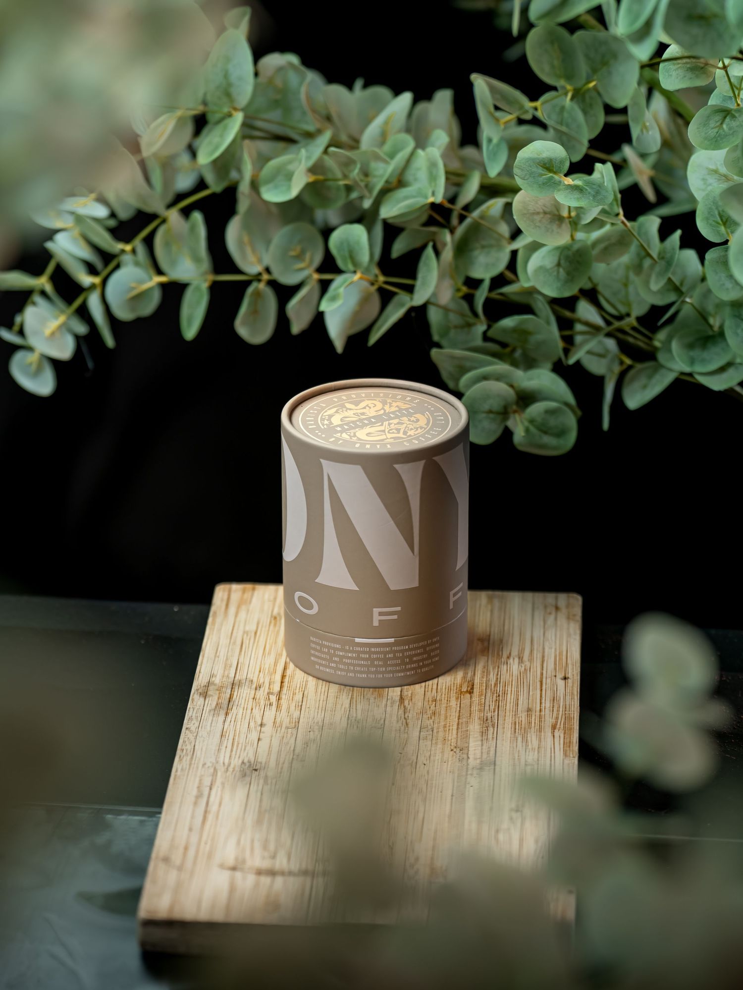 A cylindrical kraft paper coffee canister on a wooden board, surrounded by eucalyptus branches in soft natural light.
