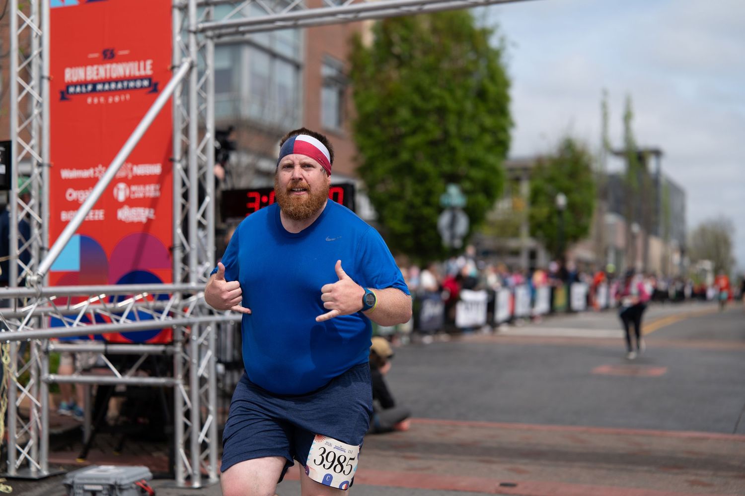 Overweight man in blue shirt and red headband running a half marathon race on a city street.