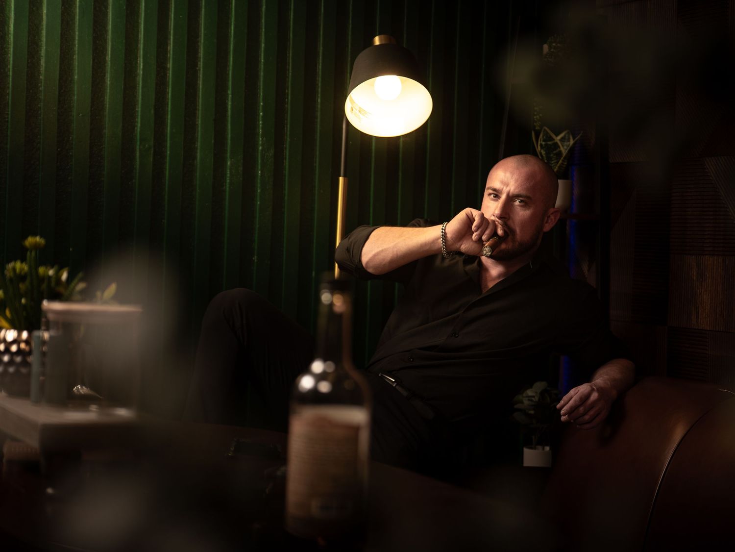 Man in black shirt sits pensively in dimly lit lounge, illuminated by warm lamp glow against green striped wall.