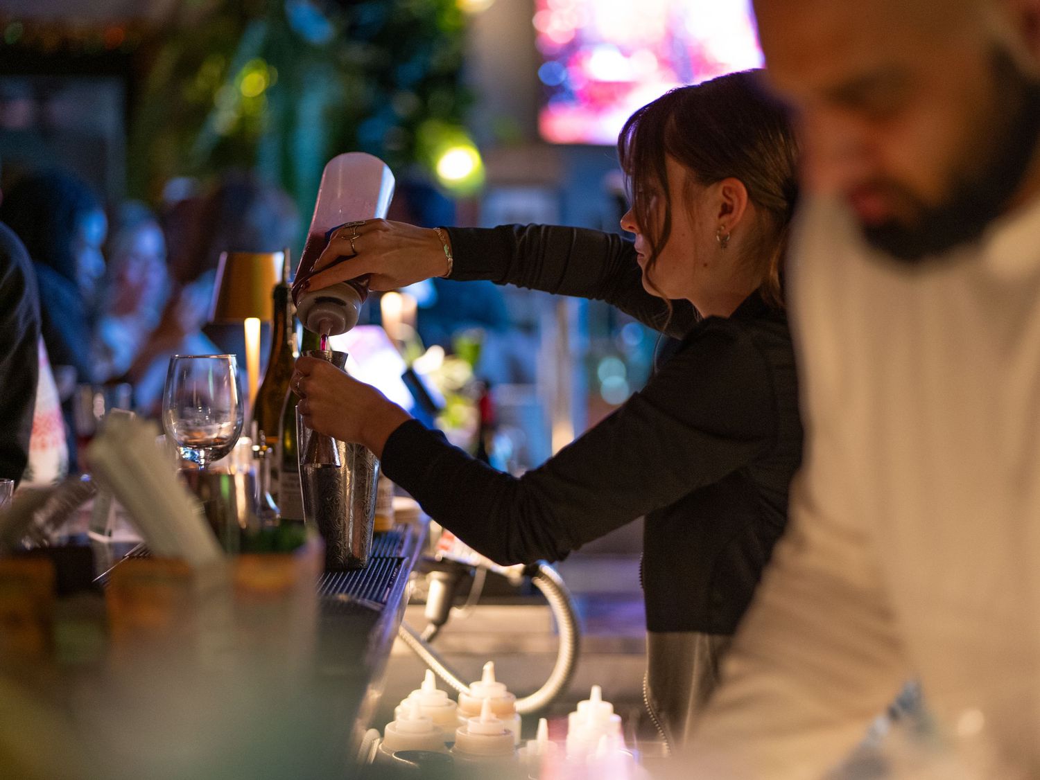 A bartender pours a cocktail into a glass at a dimly lit bar counter during evening service.