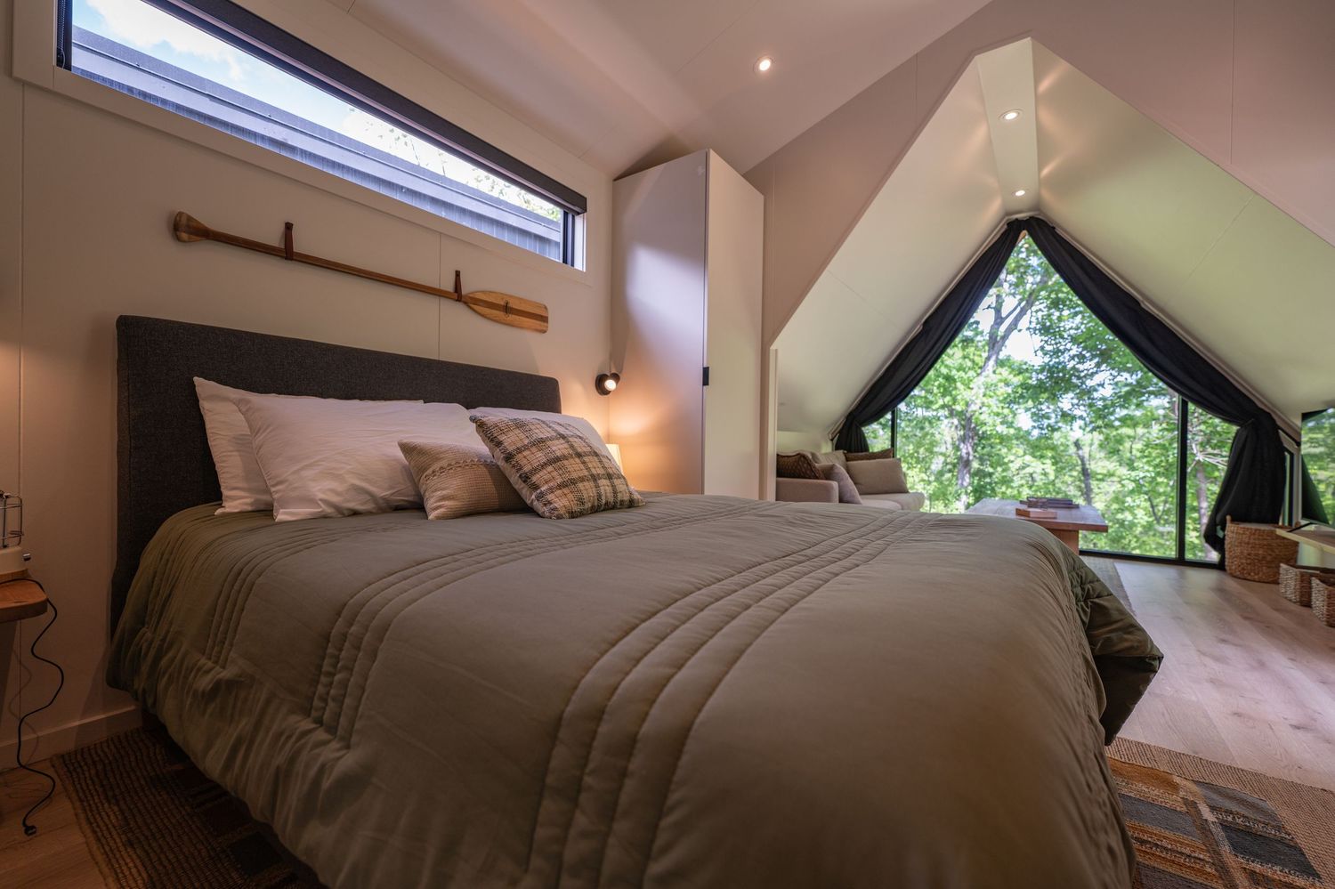 Cozy A-frame cabin bedroom with queen bed, olive bedding, wood floors, and large triangular window overlooking lush green trees.