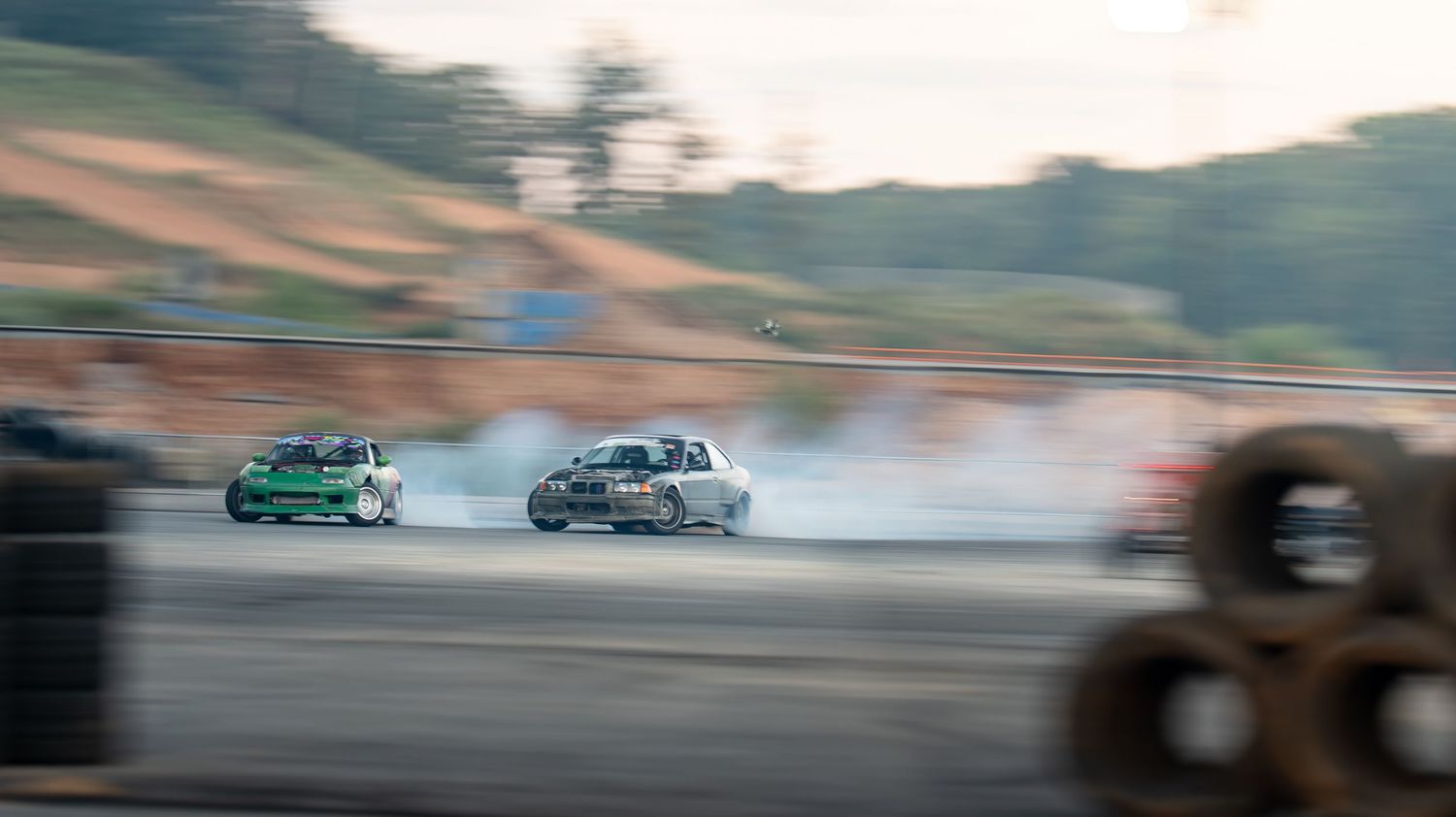 Two drift cars competing on a track with heavy tire smoke during sunset.