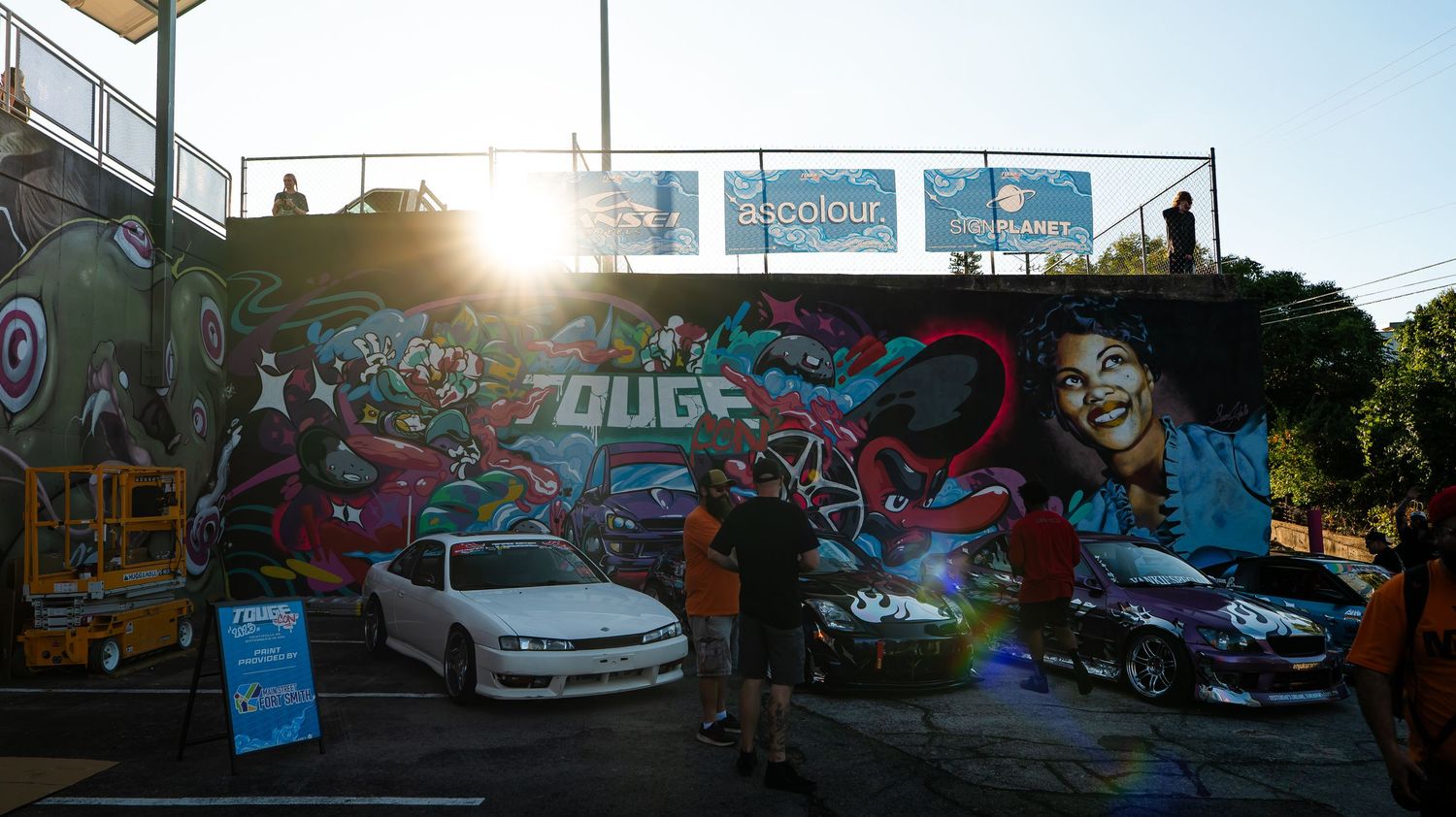 Car show panorama featuring modified vehicles against urban backdrop with street art murals.