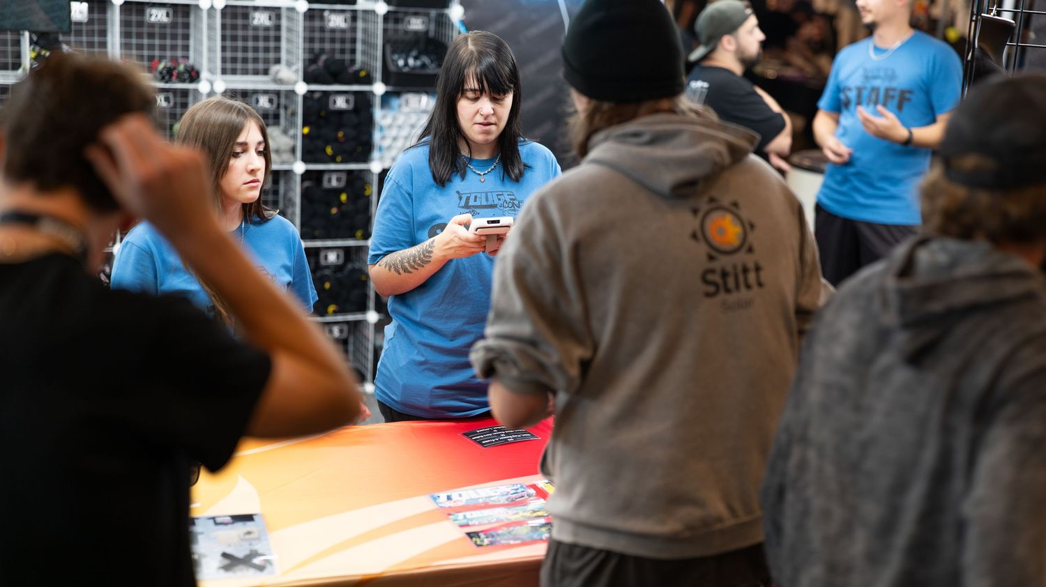 Car show attendees browse vendor displays and automotive merchandise in an indoor space.