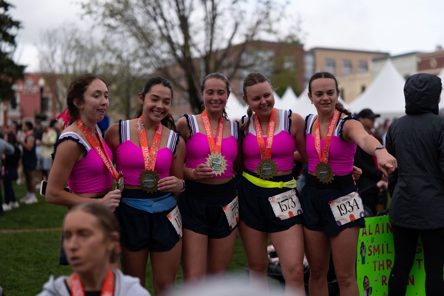 Five women in pink tops display race medals at an outdoor running event finish line celebration.