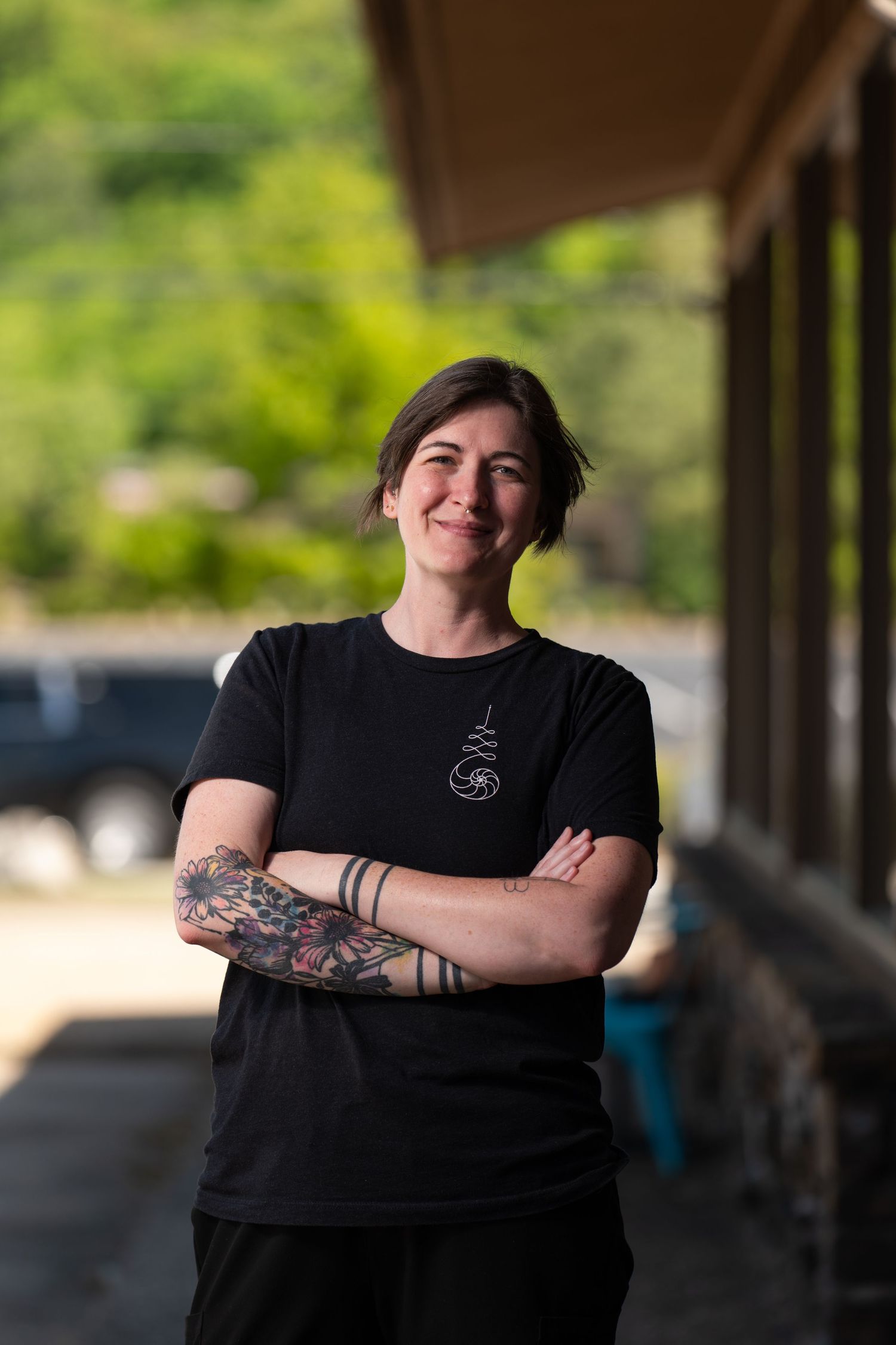 Smiling woman with tattoos and crossed arms wearing black t-shirt, standing outdoors with green trees background.