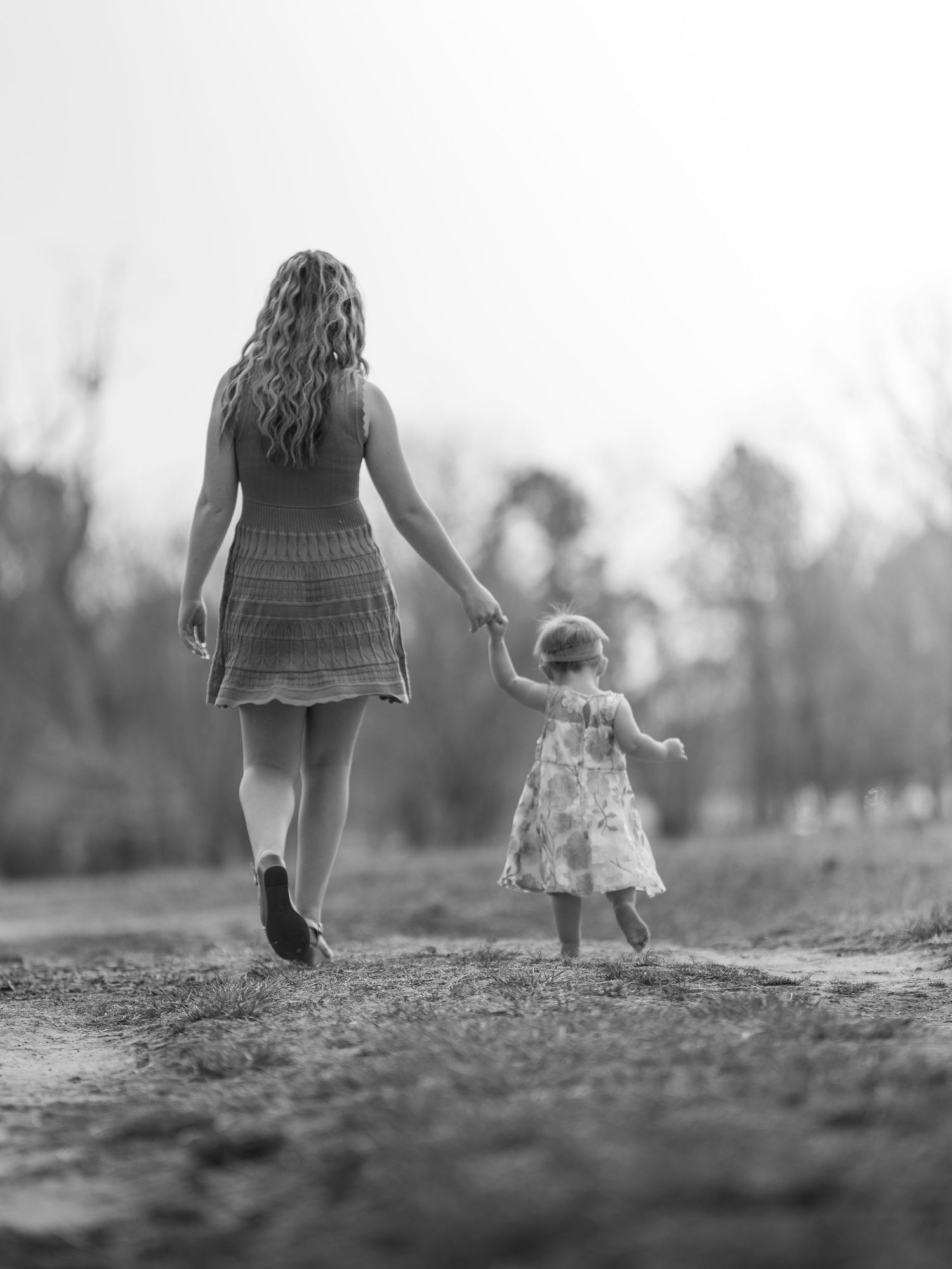 Black and white photo of a mother and young daughter holding hands while walking on a dirt path outdoors.