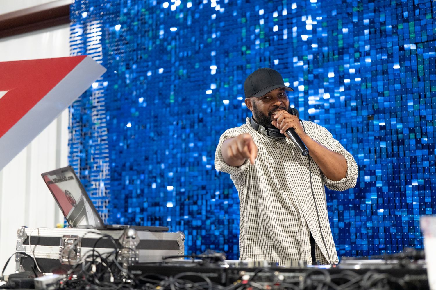 A DJ performs on stage with a microphone, pointing at the crowd in front of a sparkling blue sequin backdrop.