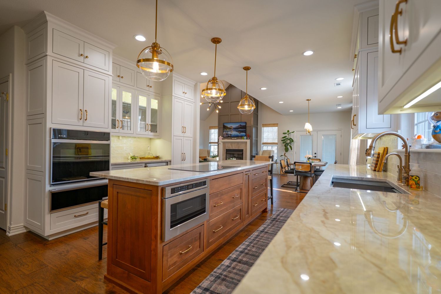Elegant kitchen with white cabinets, wood island, double ovens, gold pendant lights, and marble countertops open to living area.