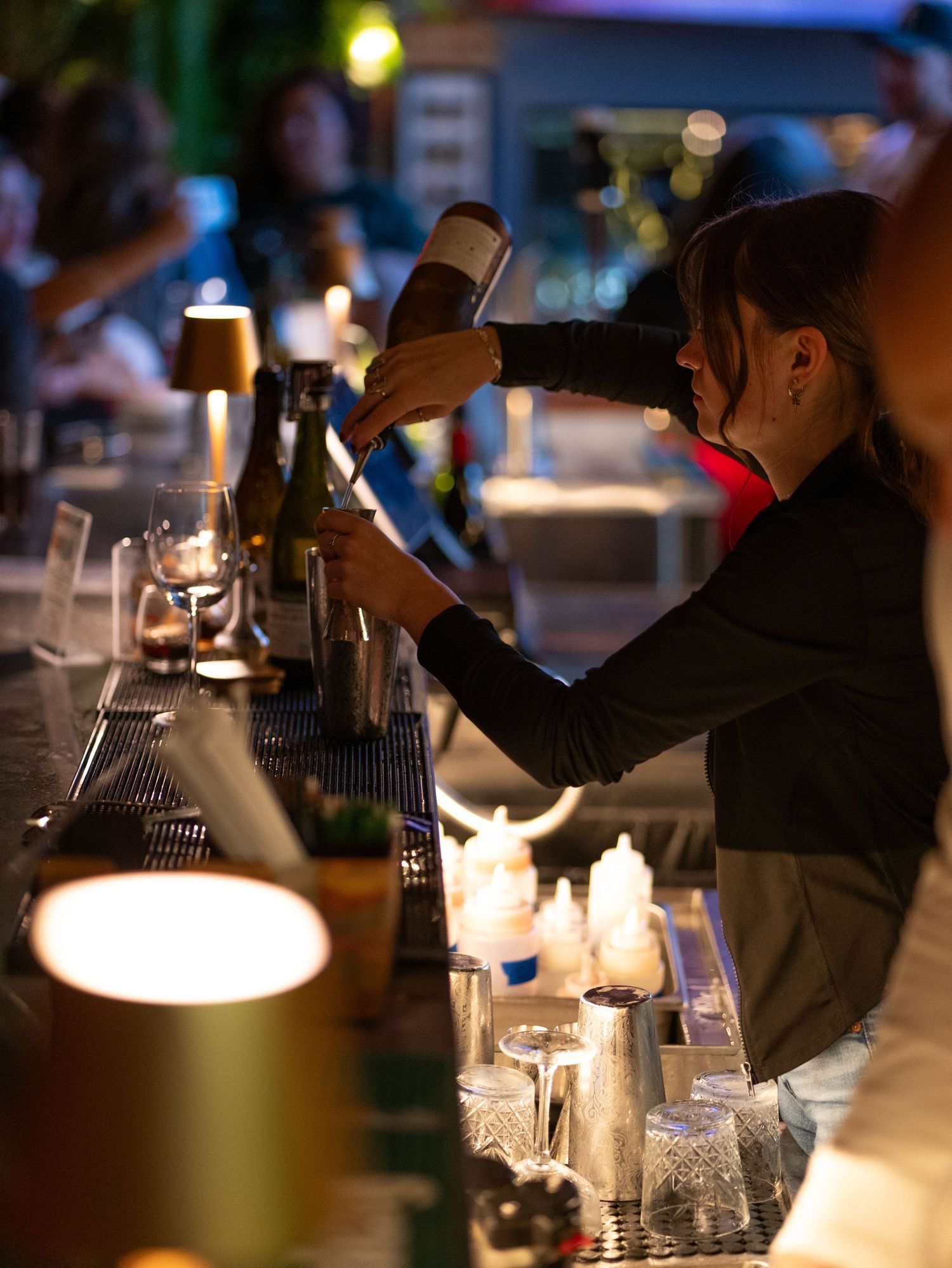 A dimly lit bar scene with multiple white desk lamps illuminating the counter space where bartenders work.
