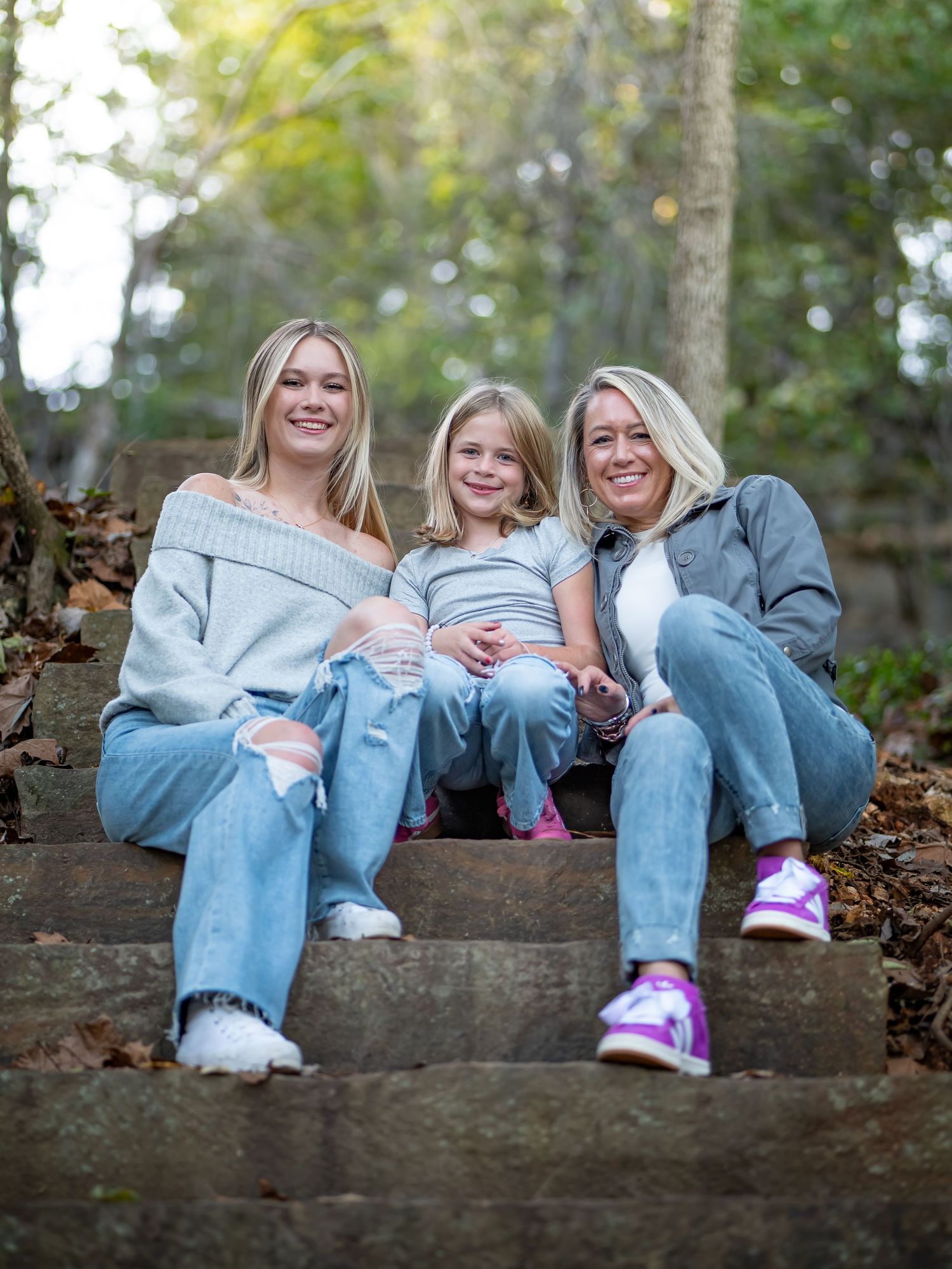 Three people sitting together on outdoor stone steps wearing casual denim and sweaters with autumn leaves around them.