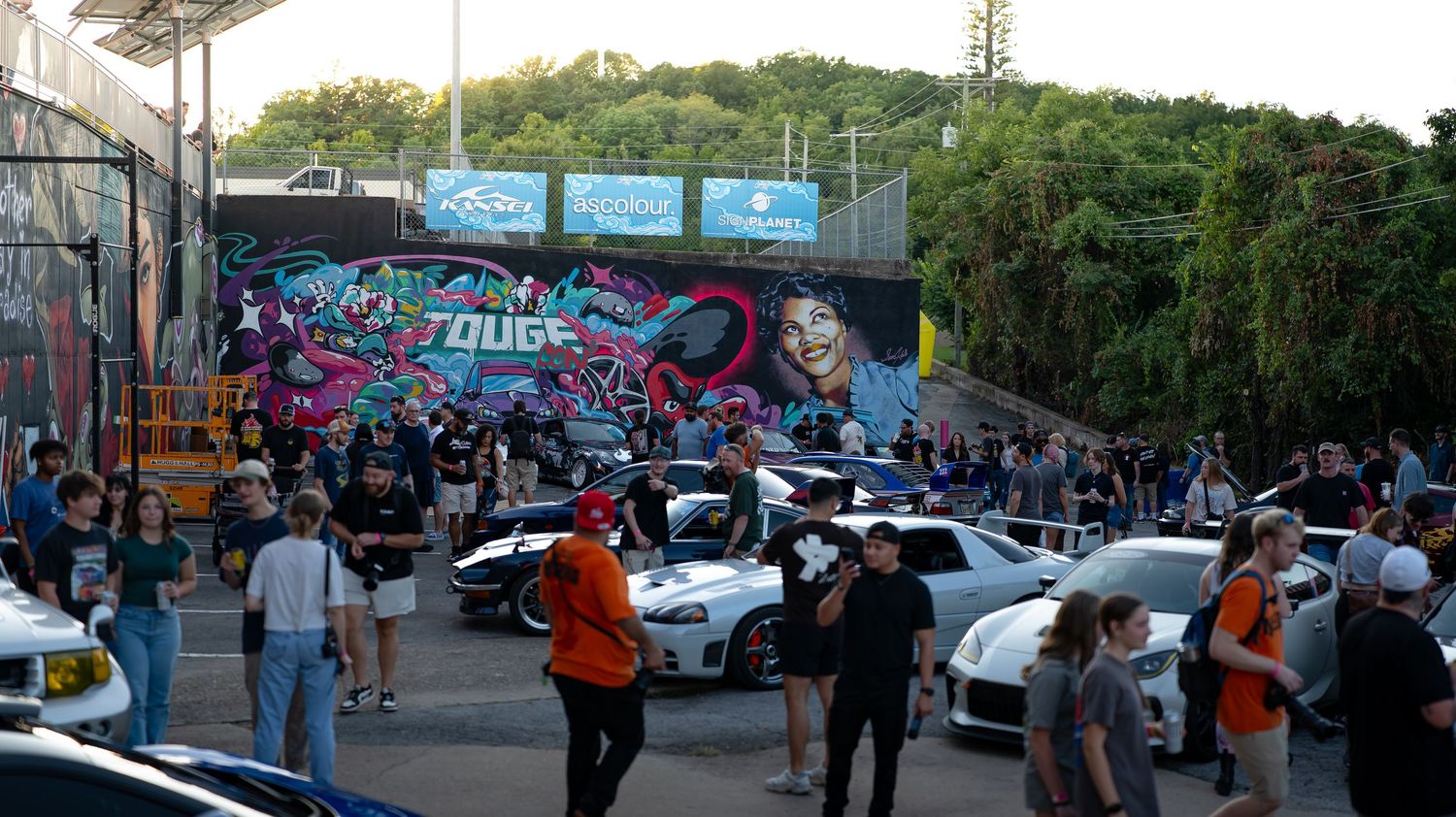 Wide panoramic view of an outdoor event with cars and people gathering in a parking area.