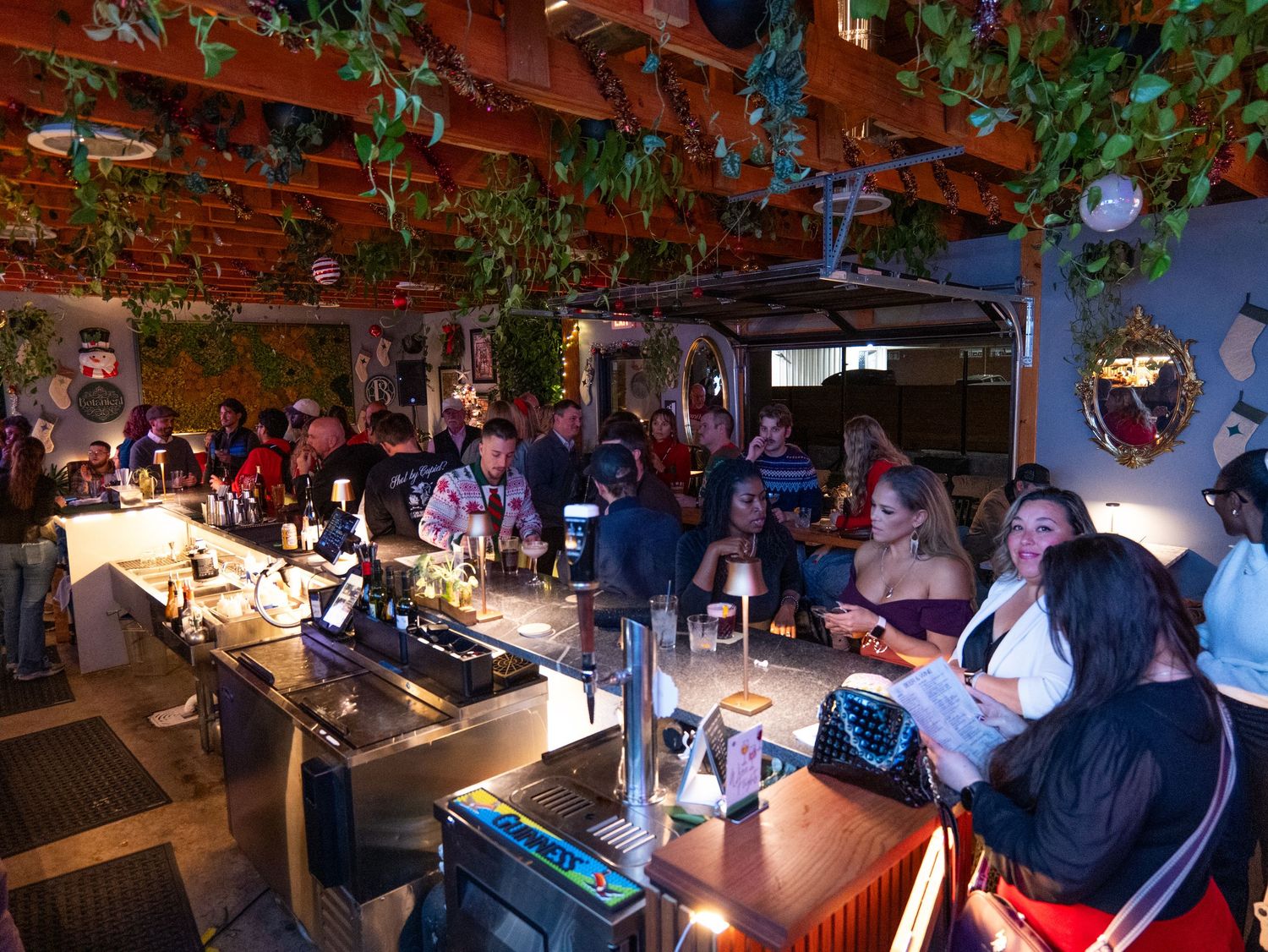 People enjoying drinks and socializing at a dimly lit bar with hanging plants and decorative lighting.