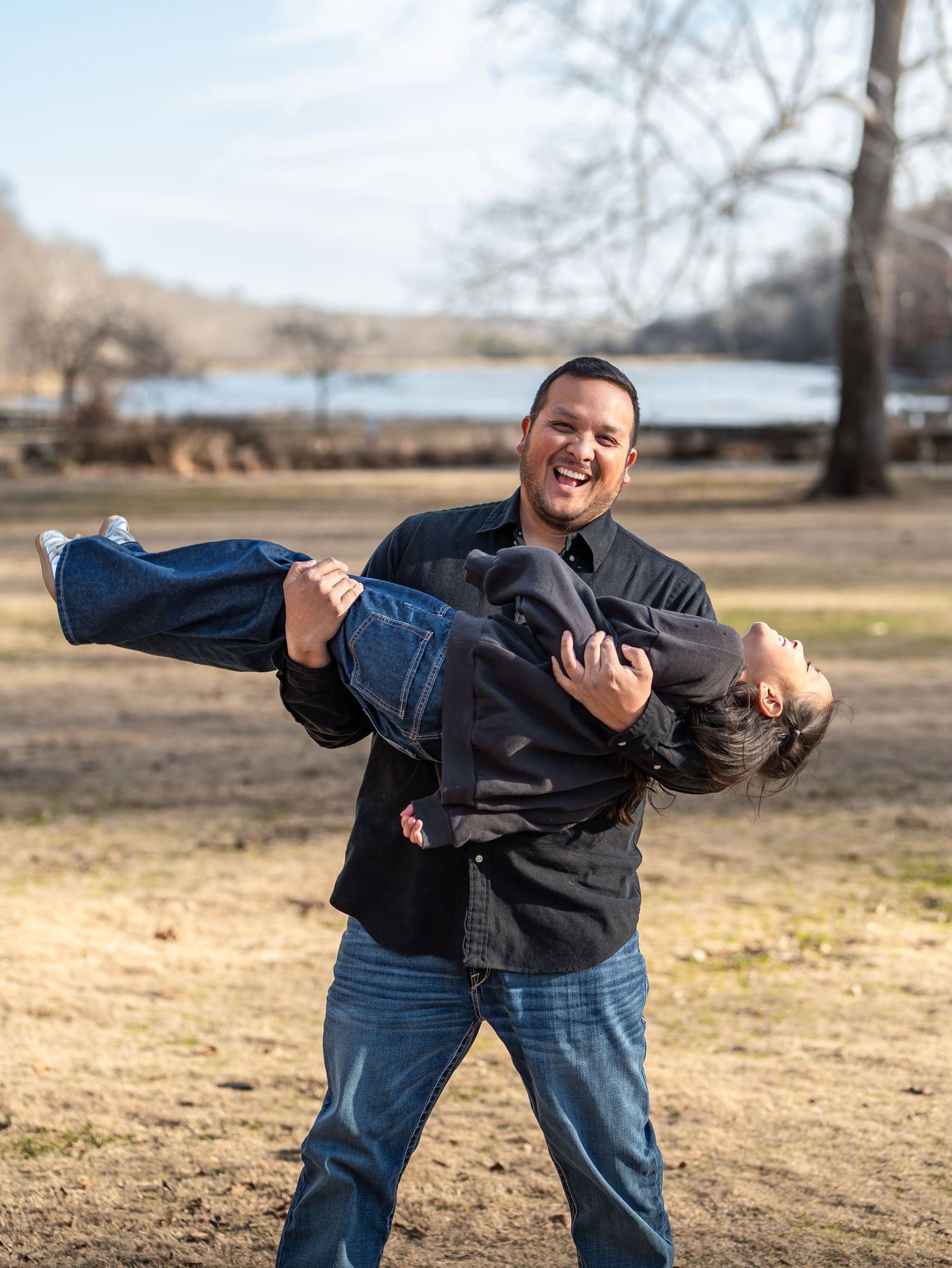 Two people share a playful moment outdoors by a lake on a winter day, with one person carrying the other.