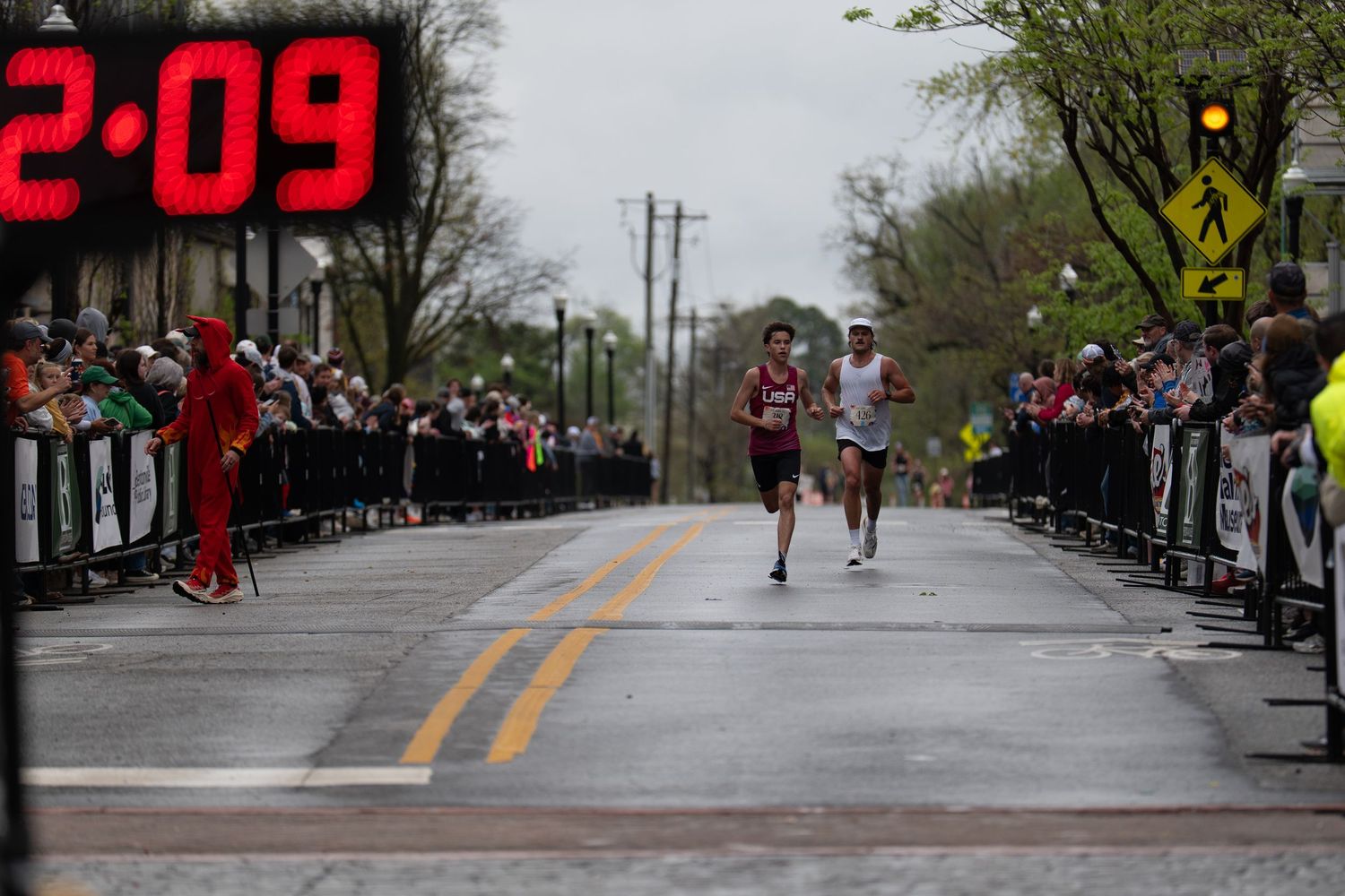 Two runners race toward the finish line at a road marathon, with a digital clock showing 2:09 and spectators lining the wet street.