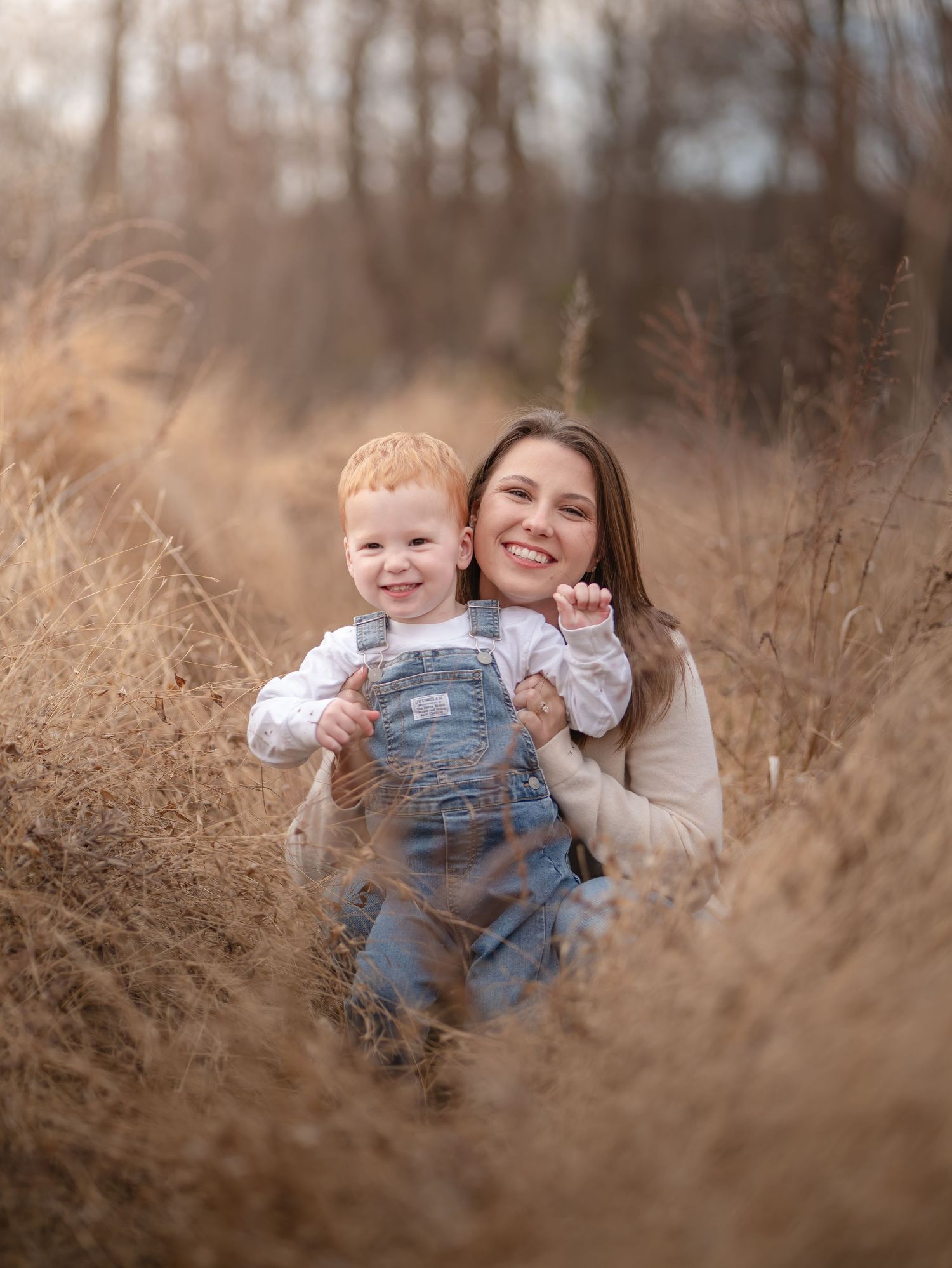 A mother and young child share joyful moments together in a field of tall brown grass during golden hour.